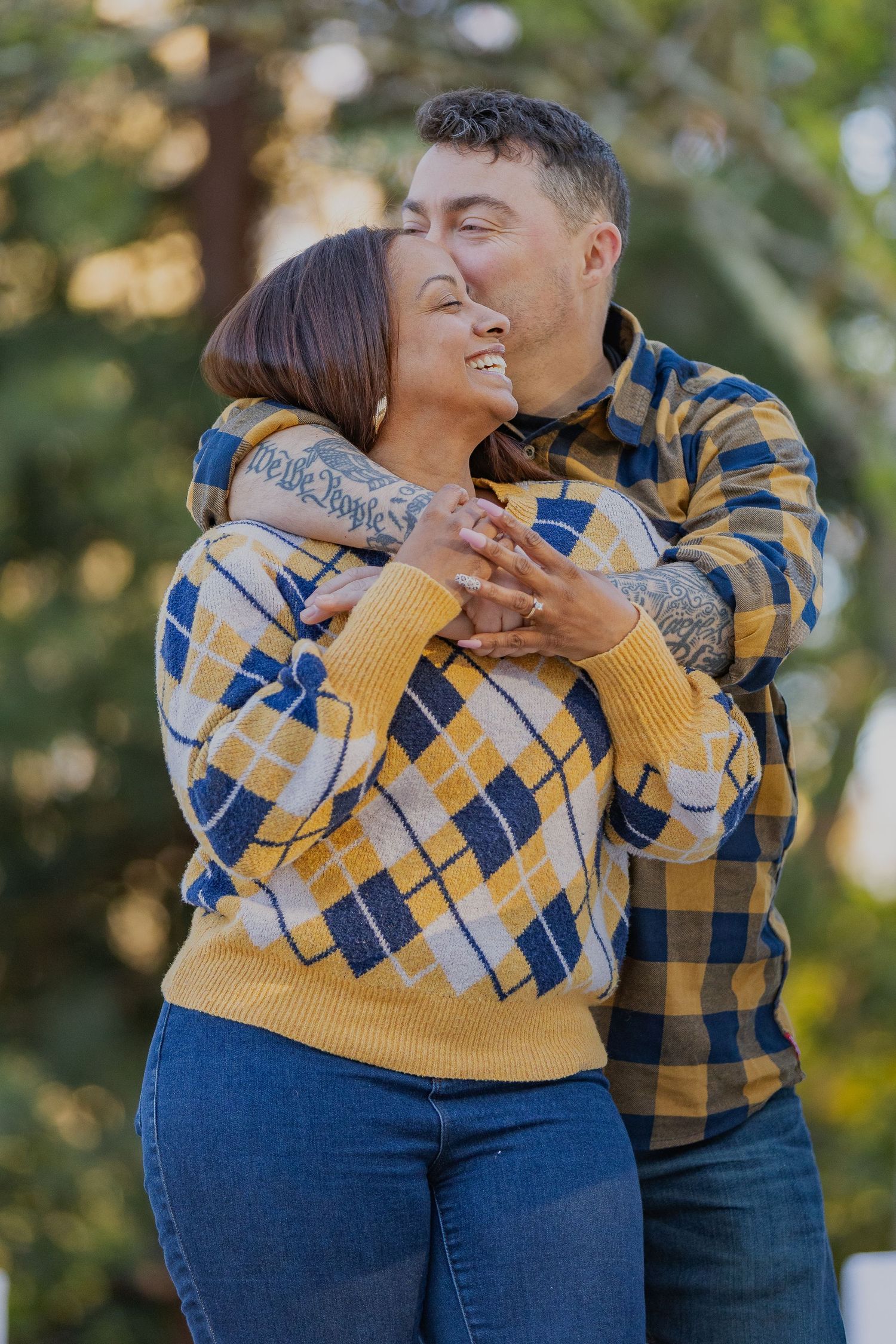 A loving couple embraces outdoors wearing matching plaid patterns in yellow and blue with tattoos visible on their arms.
