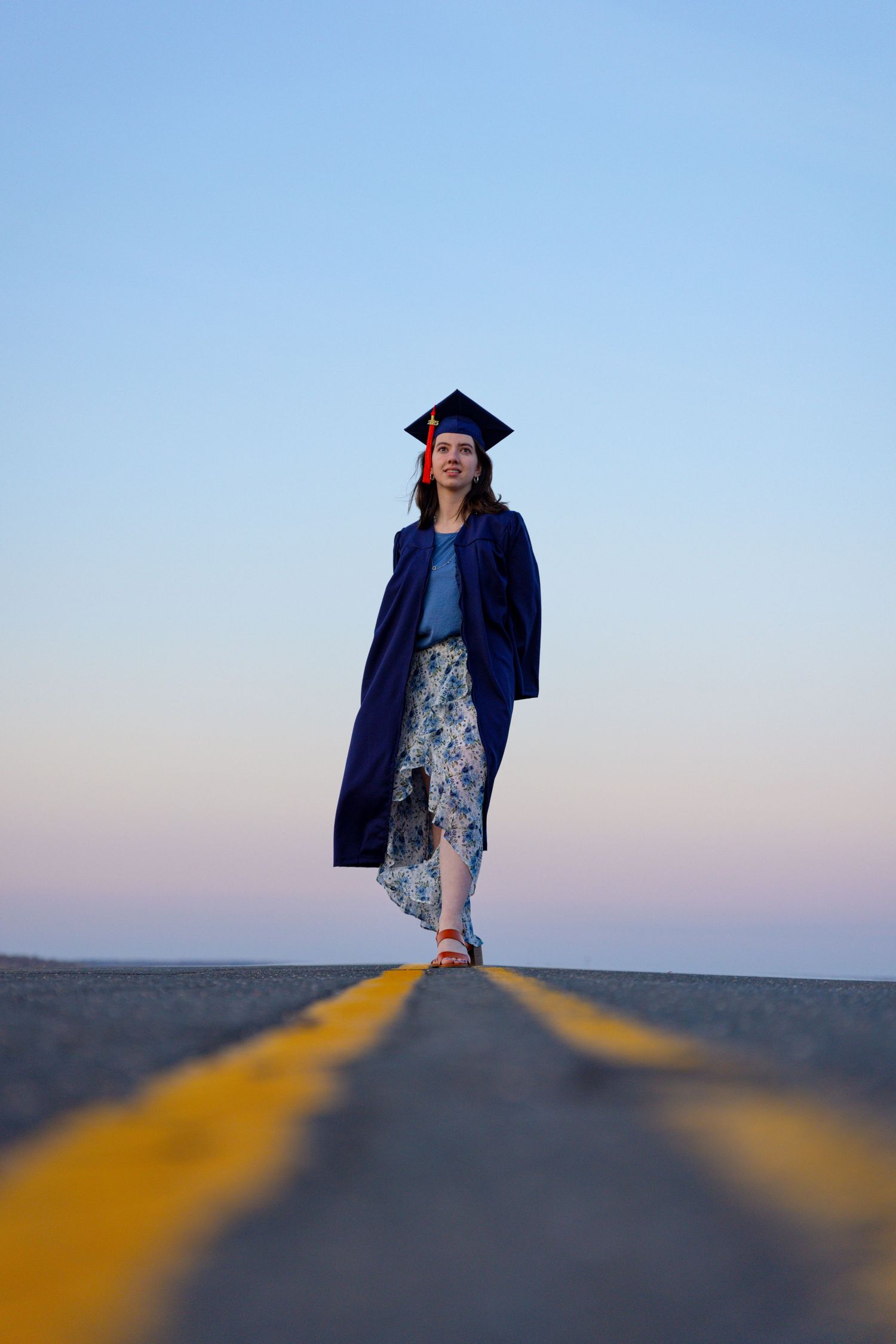Graduate in navy blue cap and gown stands on yellow road lines at sunset.