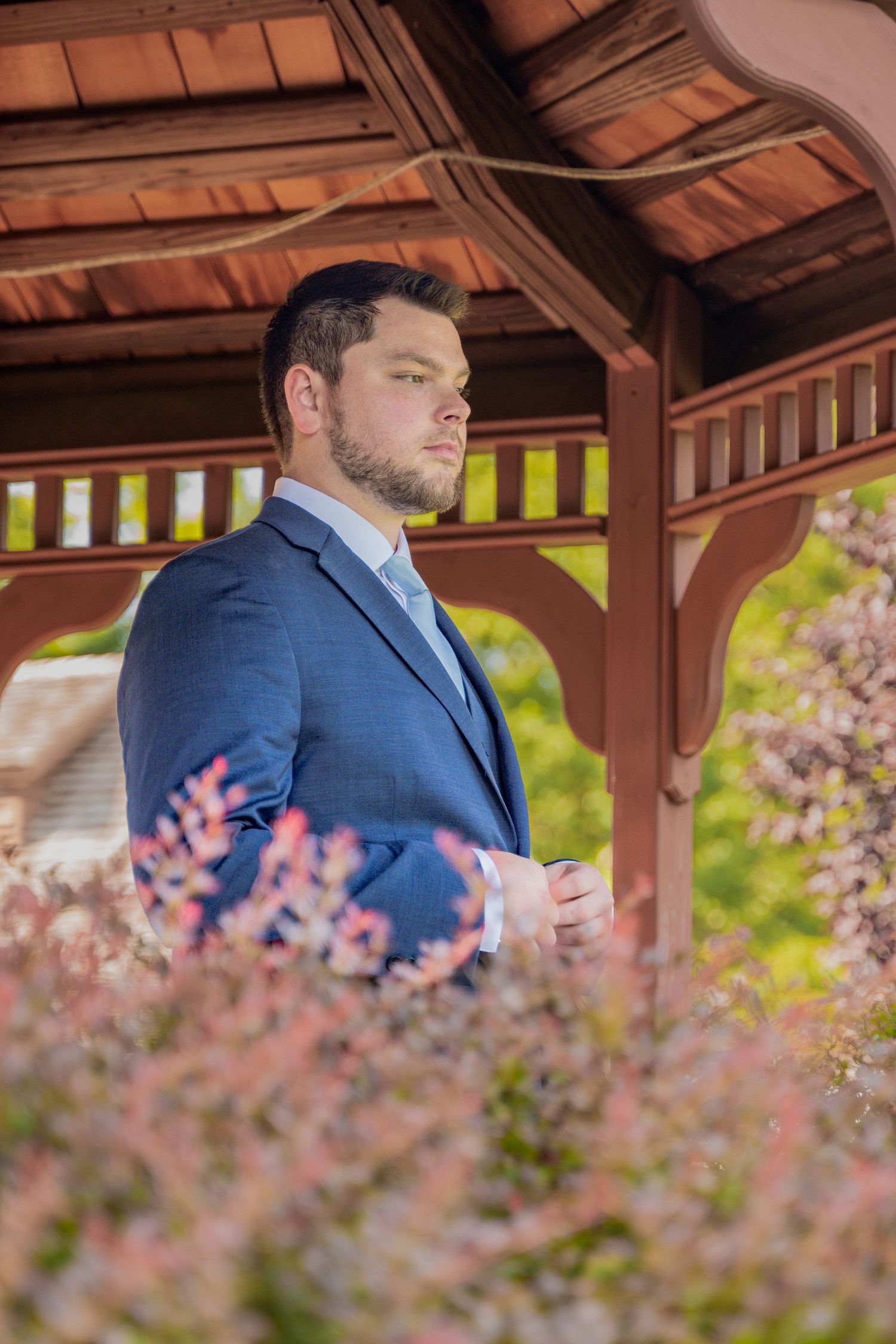 Elegant groom in blue suit poses in wooden gazebo surrounded by pink flowers.