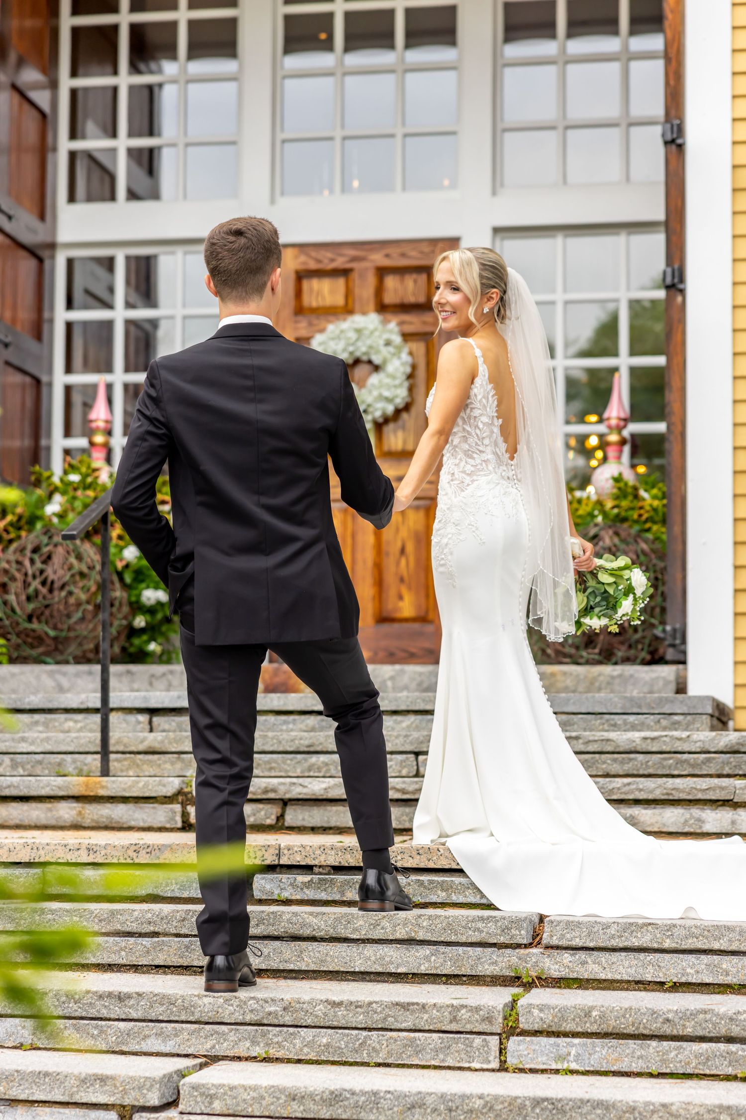 Couple in wedding attire ascending stone steps in front of a building with flowering planters.