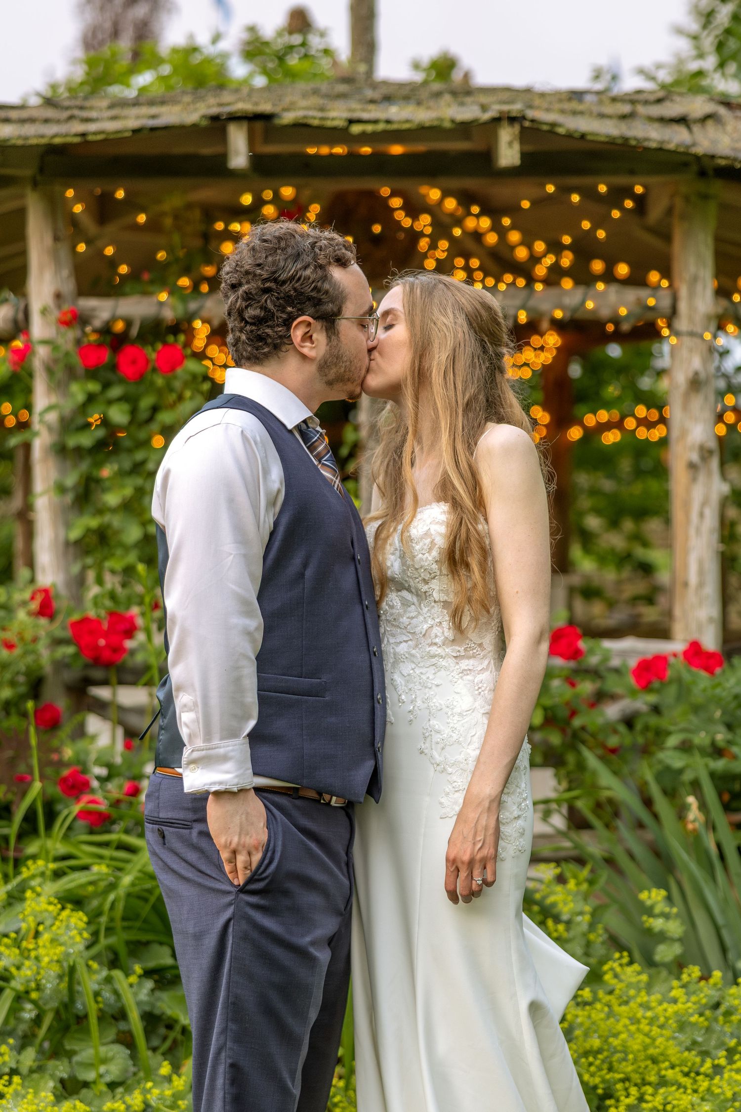 A romantic wedding scene in a garden with red roses and twinkling lights under a rustic wooden pergola.