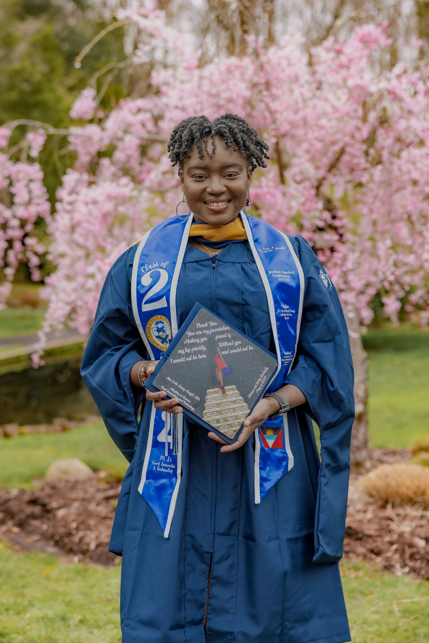 Graduate holding decorated cap stands in blue graduation gown and stole beneath blooming pink cherry blossoms.