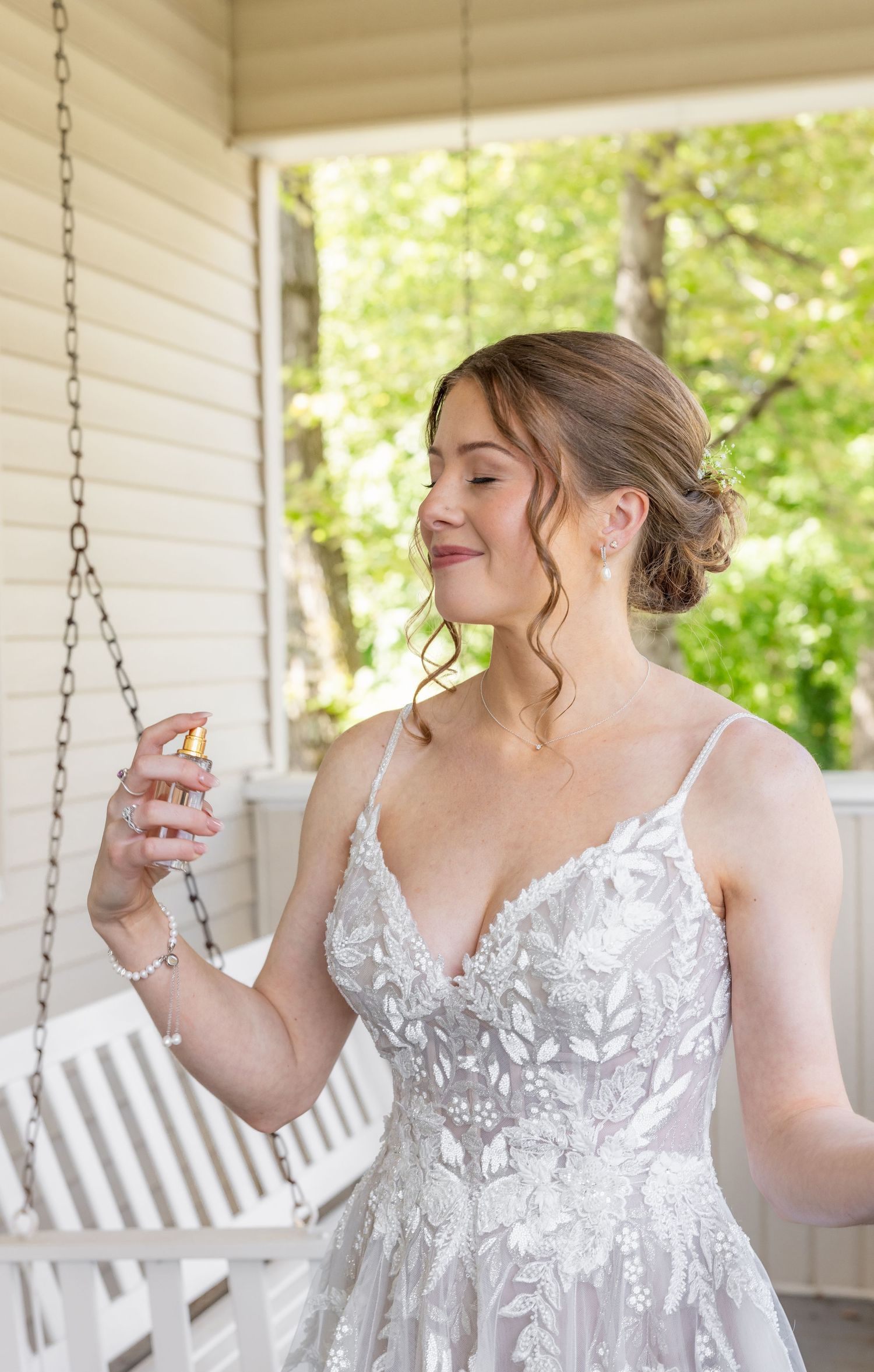 A bride in lace dress sprays perfume on her wedding day on a rustic porch swing.