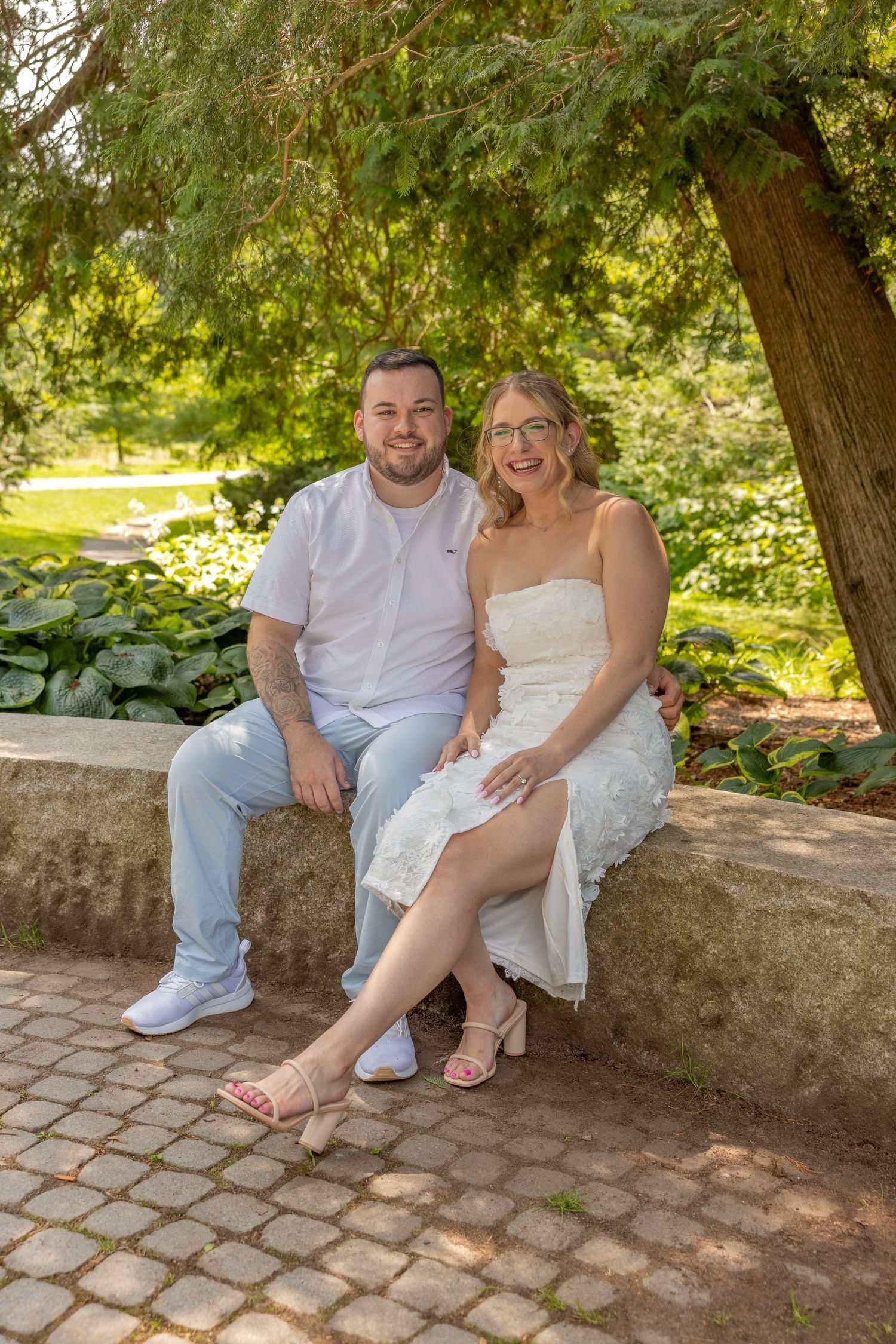 A couple in summer attire sitting together on a stone wall in a lush garden.
