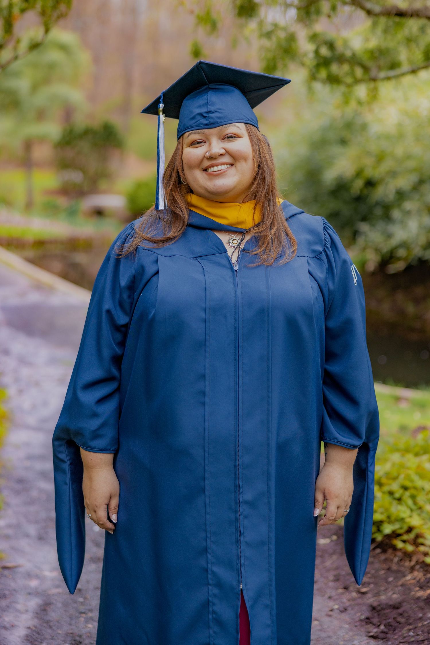 Graduate poses outdoors in traditional blue academic regalia with yellow accent scarf.