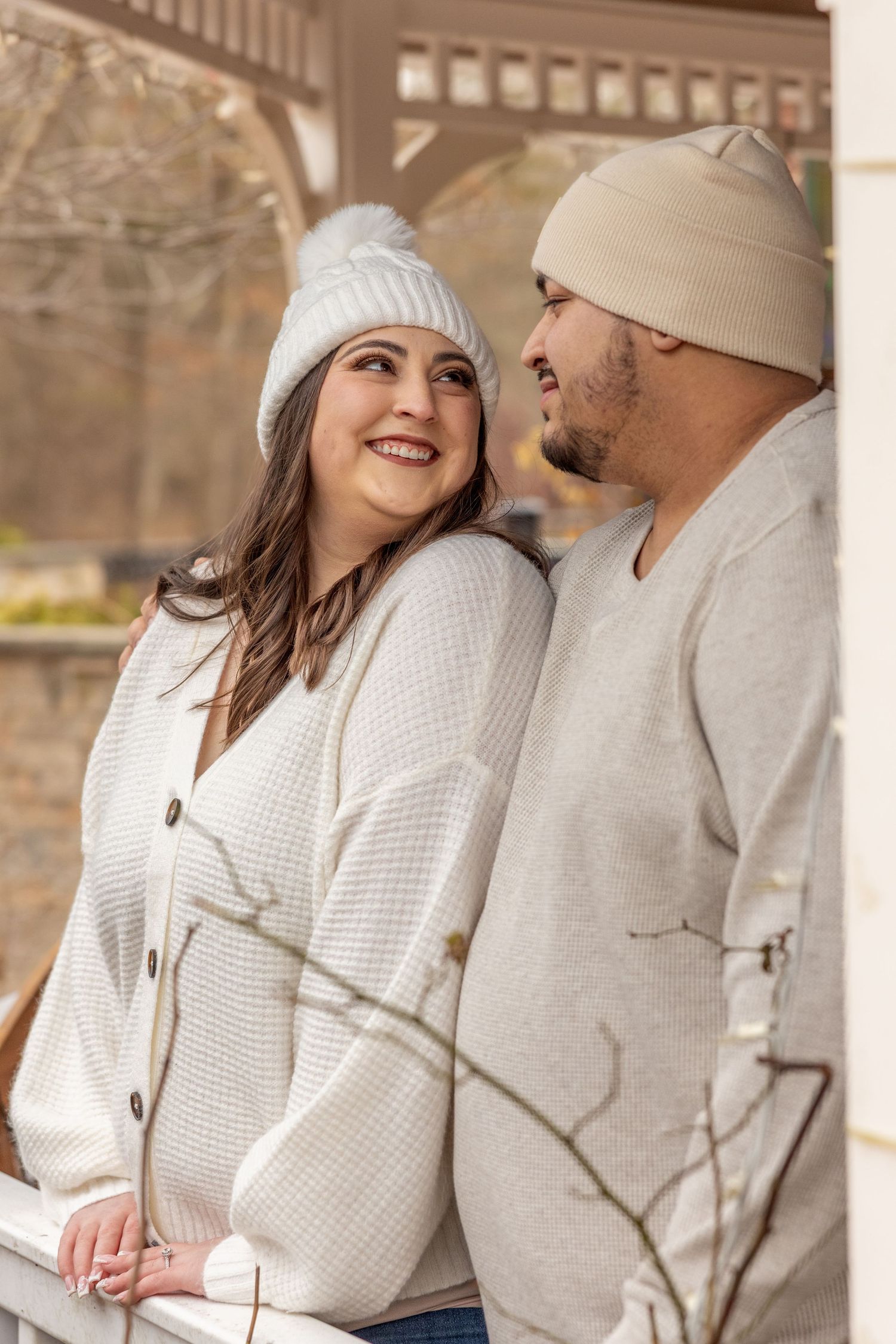 A couple wearing winter hats and white sweaters smiles at each other on a covered porch.