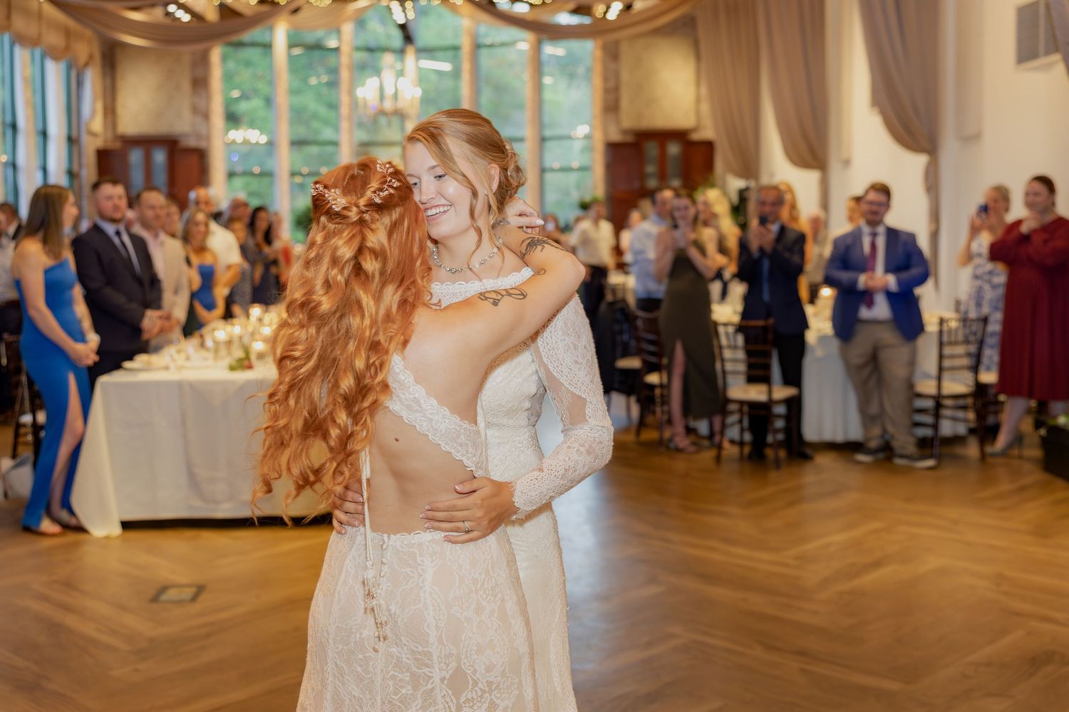 Two people share an intimate dance moment on a wooden floor at an elegant reception venue with guests looking on.