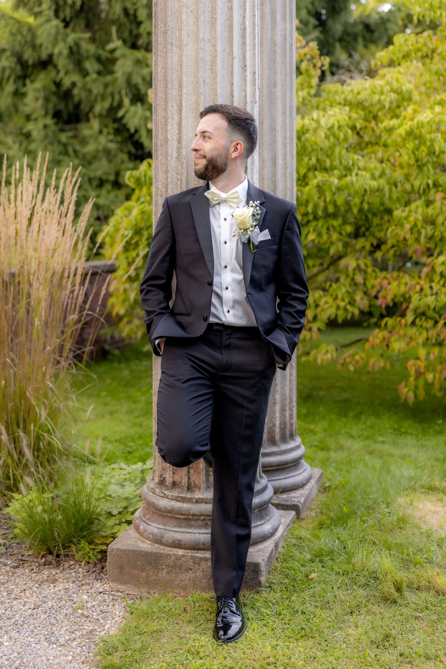 Elegant groom in black tuxedo poses against classical stone column in garden setting.