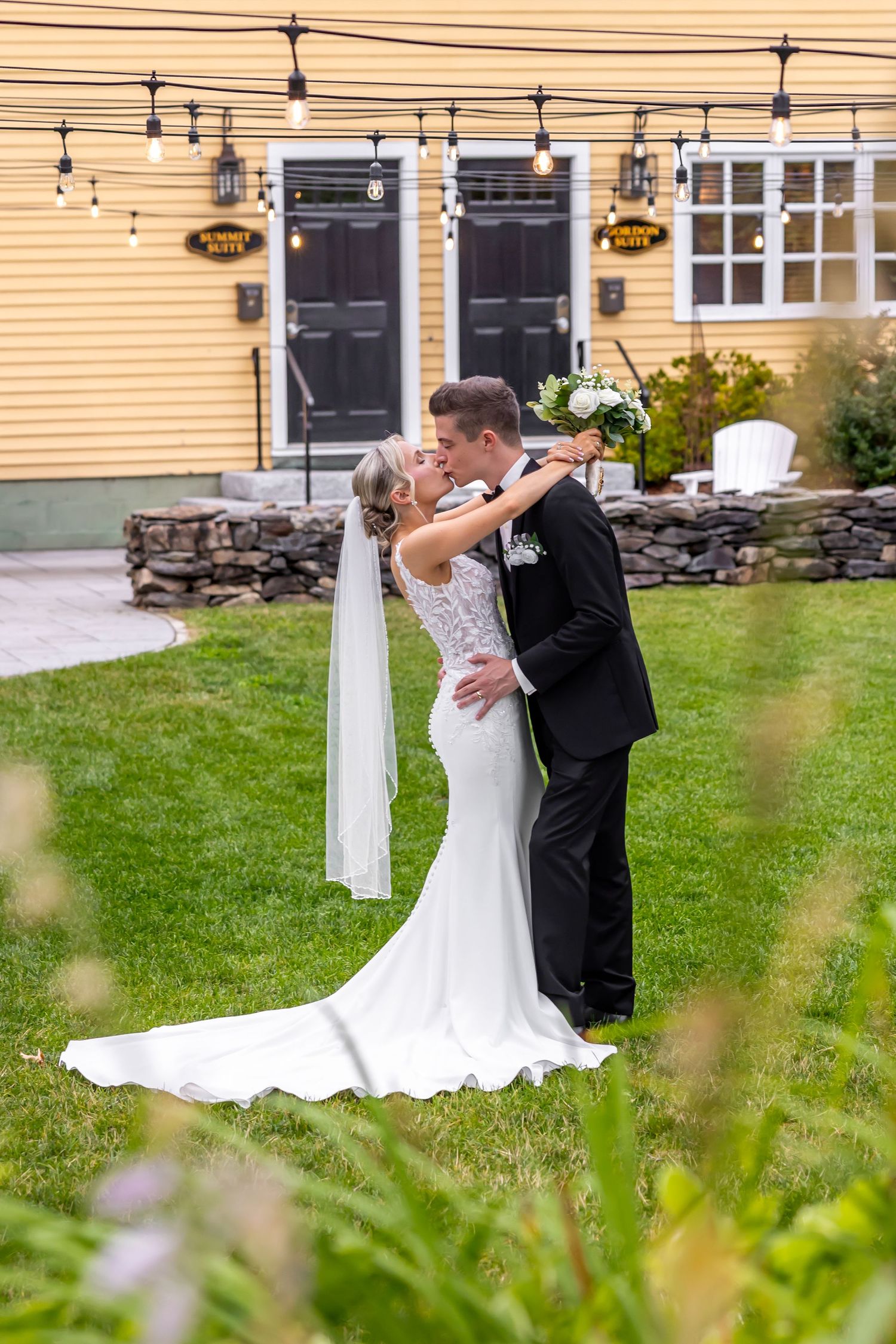 Bride and groom share intimate kiss in front of yellow house with string lights and stone wall.
