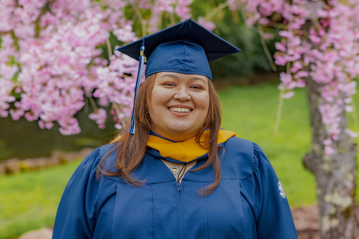 Graduate in blue cap and gown stands smiling against pink cherry blossoms in springtime garden.