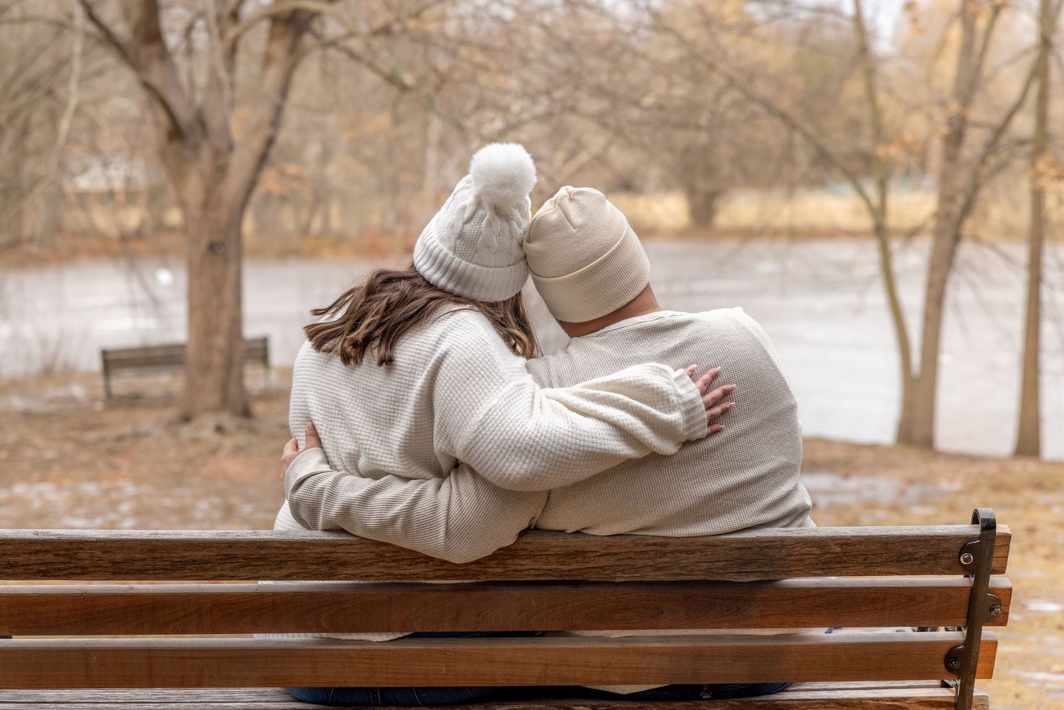 A couple in matching cream sweaters and beanies sitting close on a park bench by a lake.