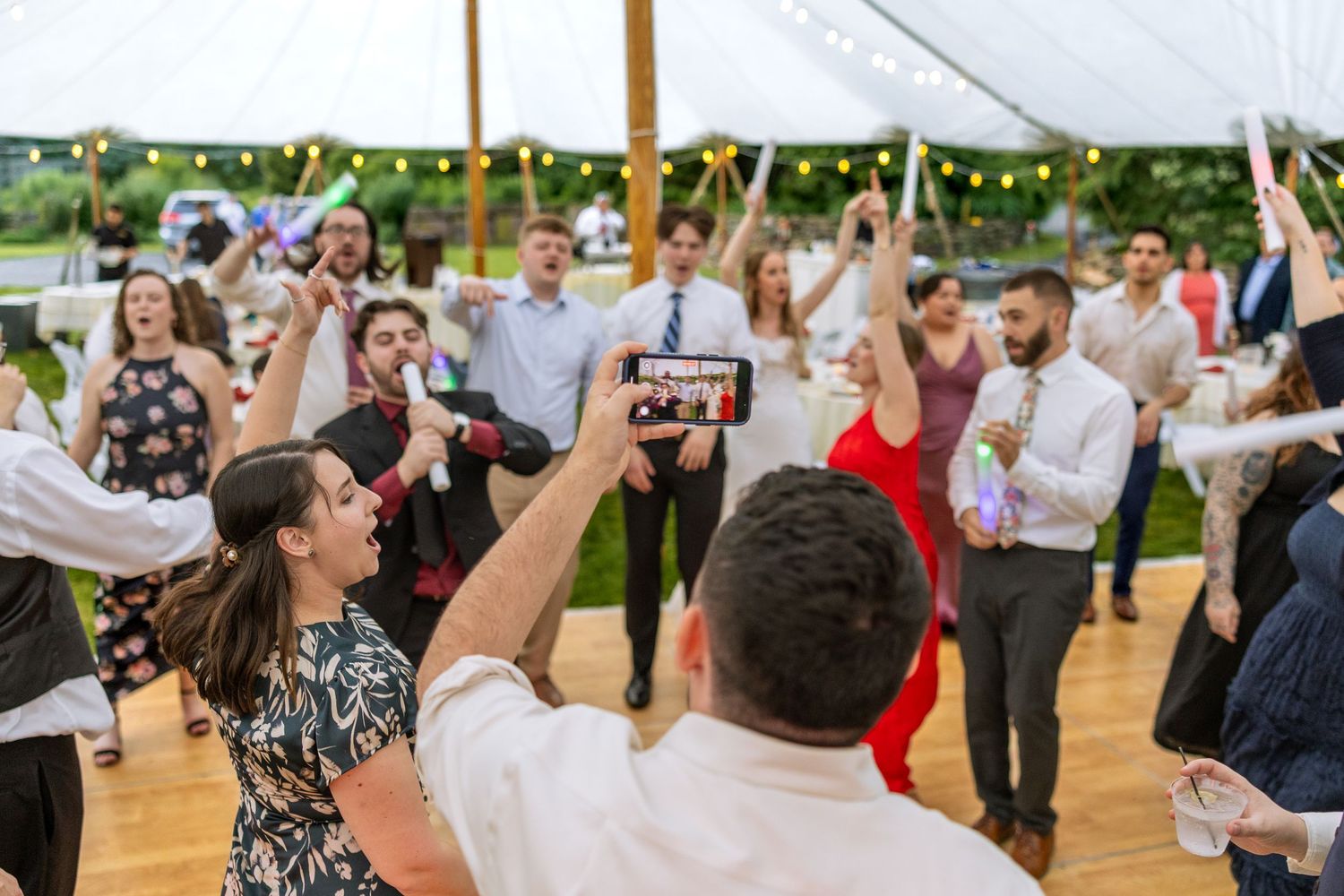 Wedding guests dance and celebrate together on a wooden dance floor under an outdoor tent during a reception celebration.