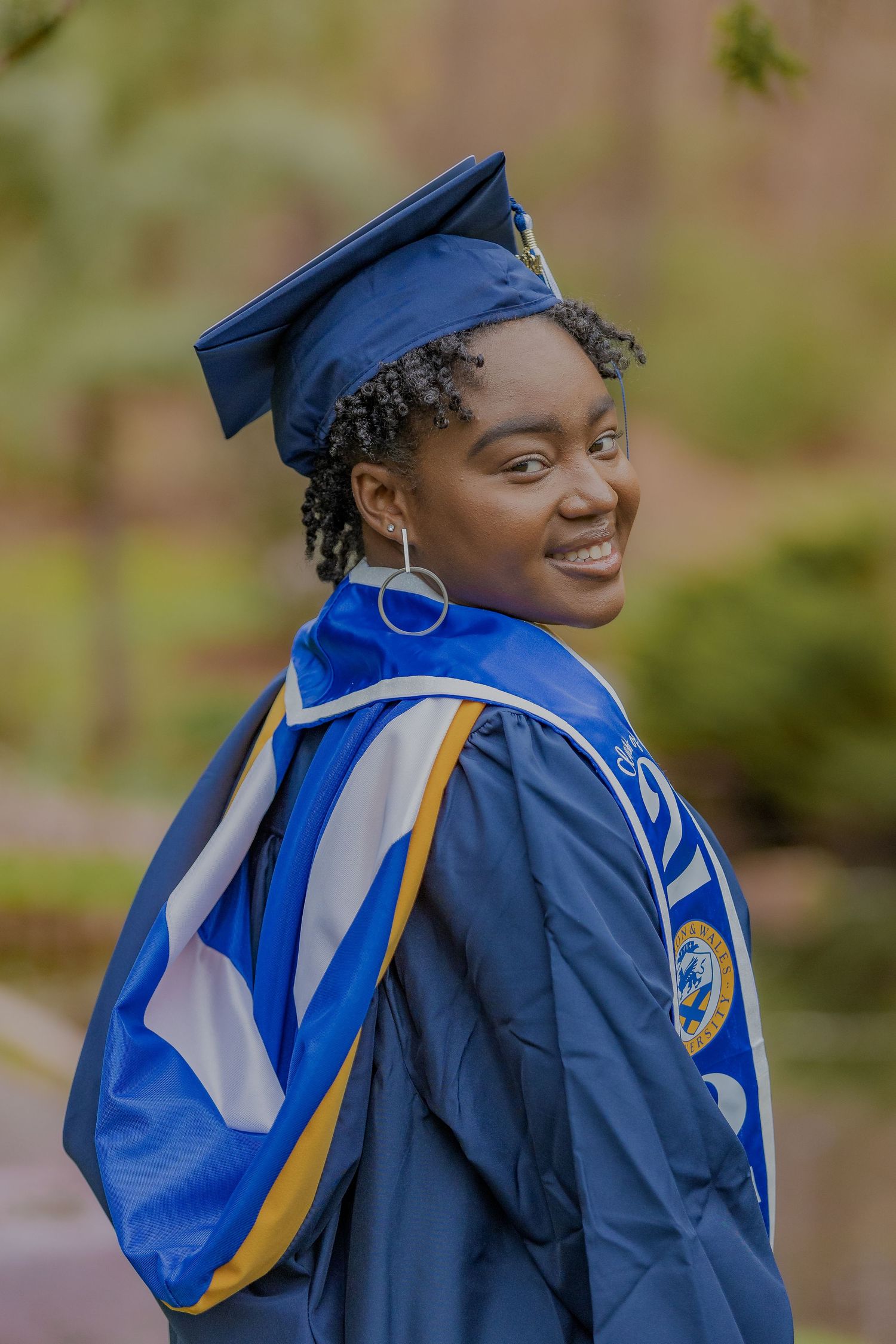 Graduate in blue academic regalia and honors cords smiling against blurred natural background.