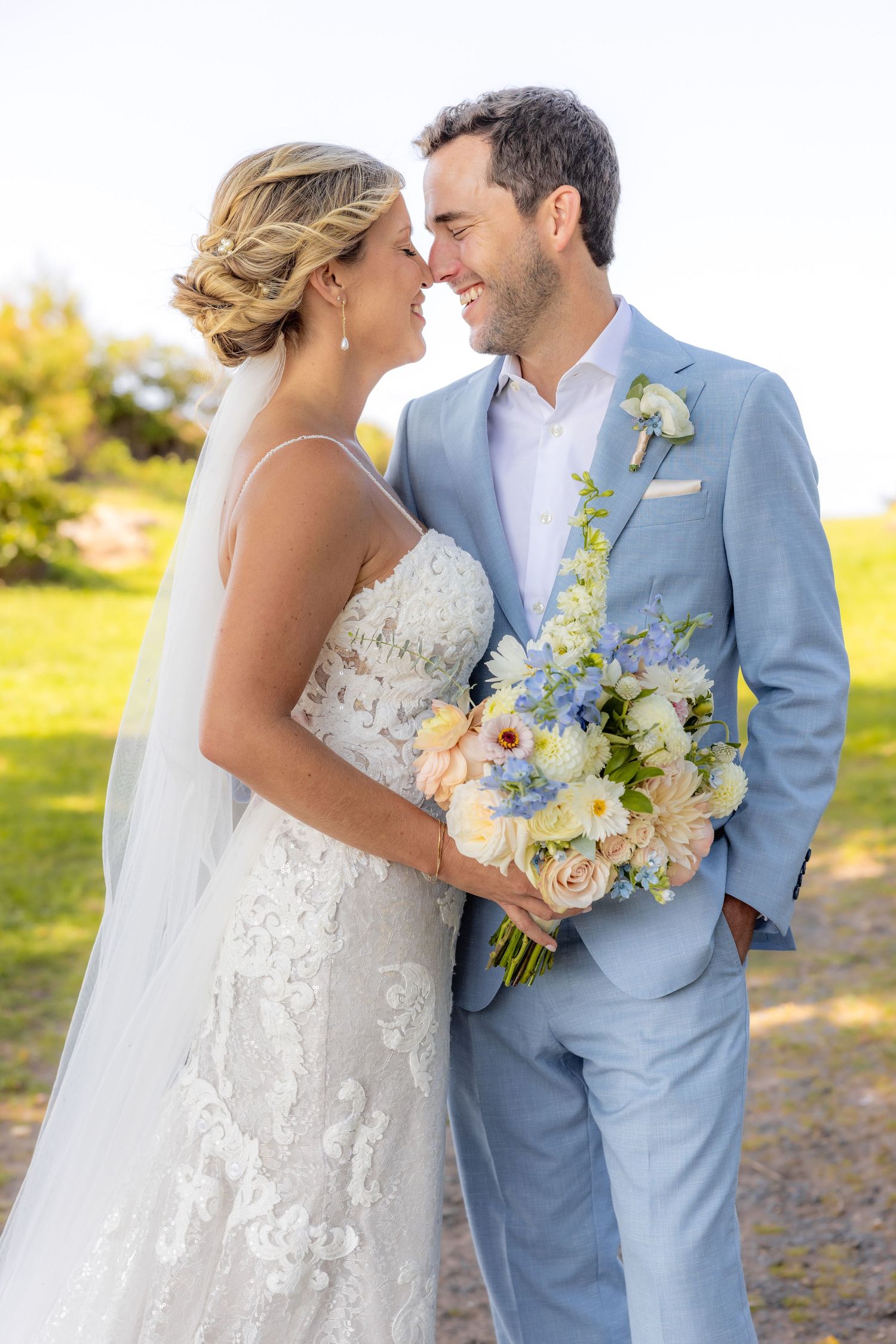 Couple in wedding attire holding delicate blue and white bouquet in sunny outdoor setting.