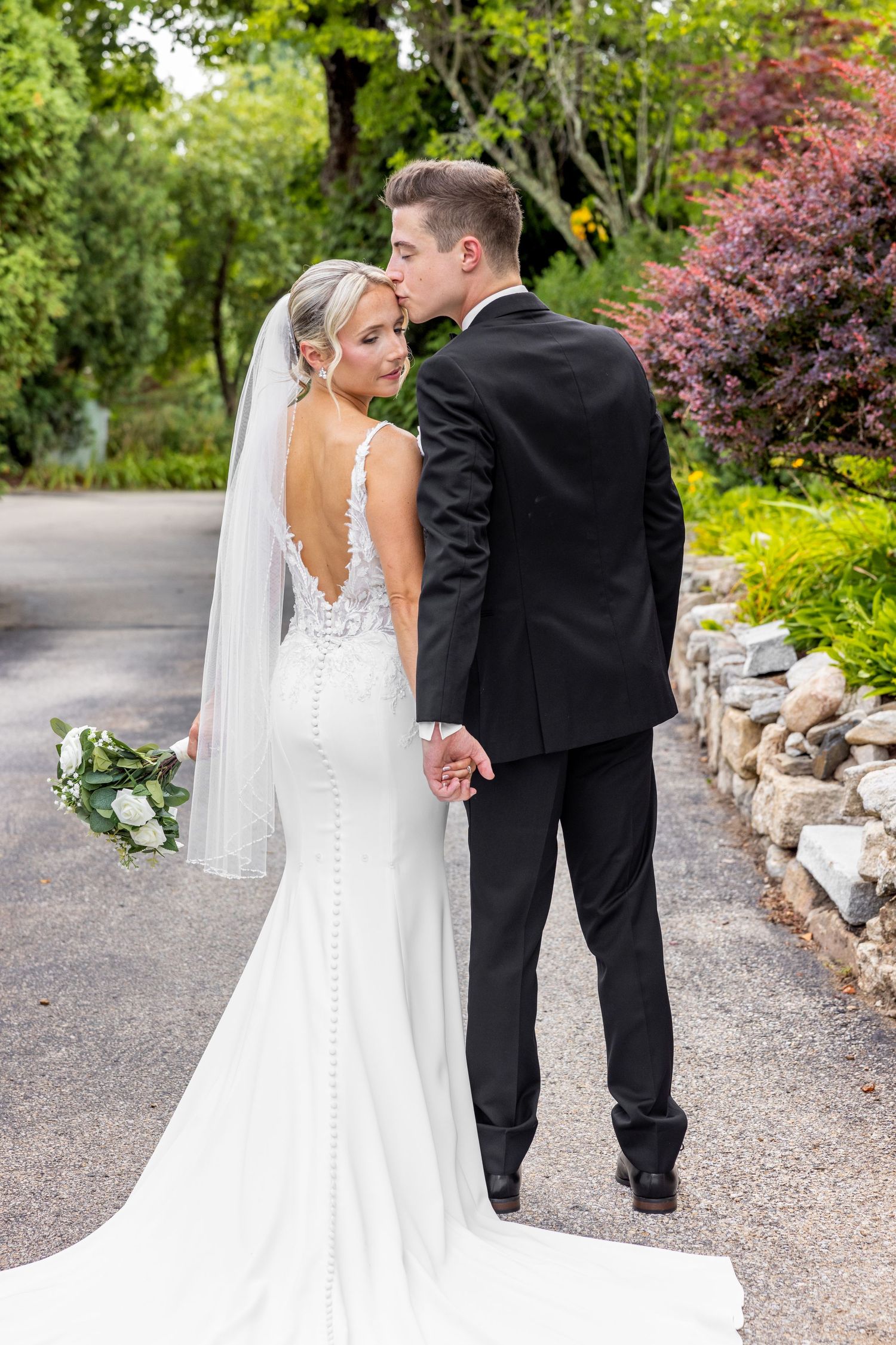 Elegant bride in fitted lace gown and groom walking hand-in-hand on garden path.