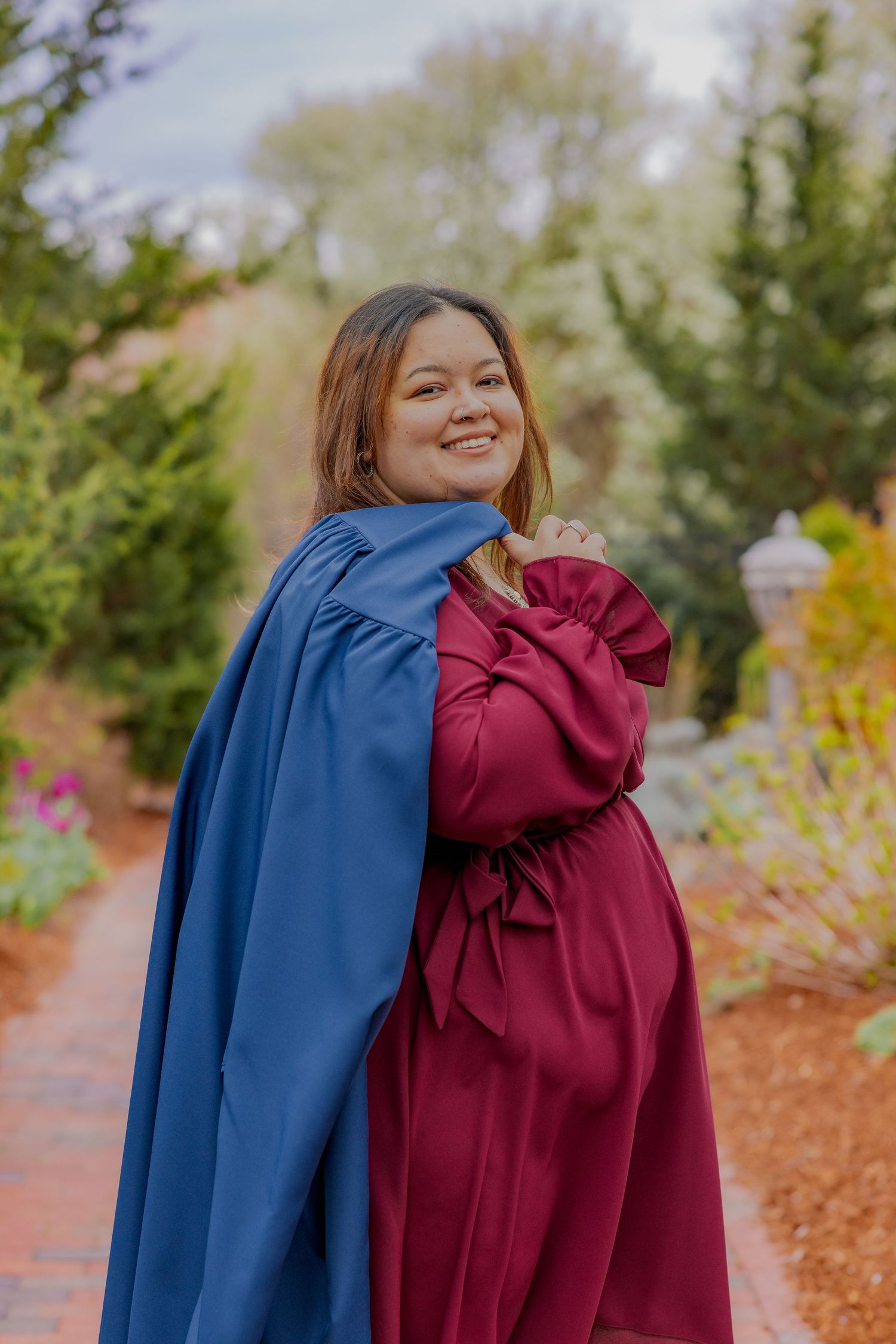 Person holds burgundy dress while wearing blue graduation gown in outdoor garden setting.