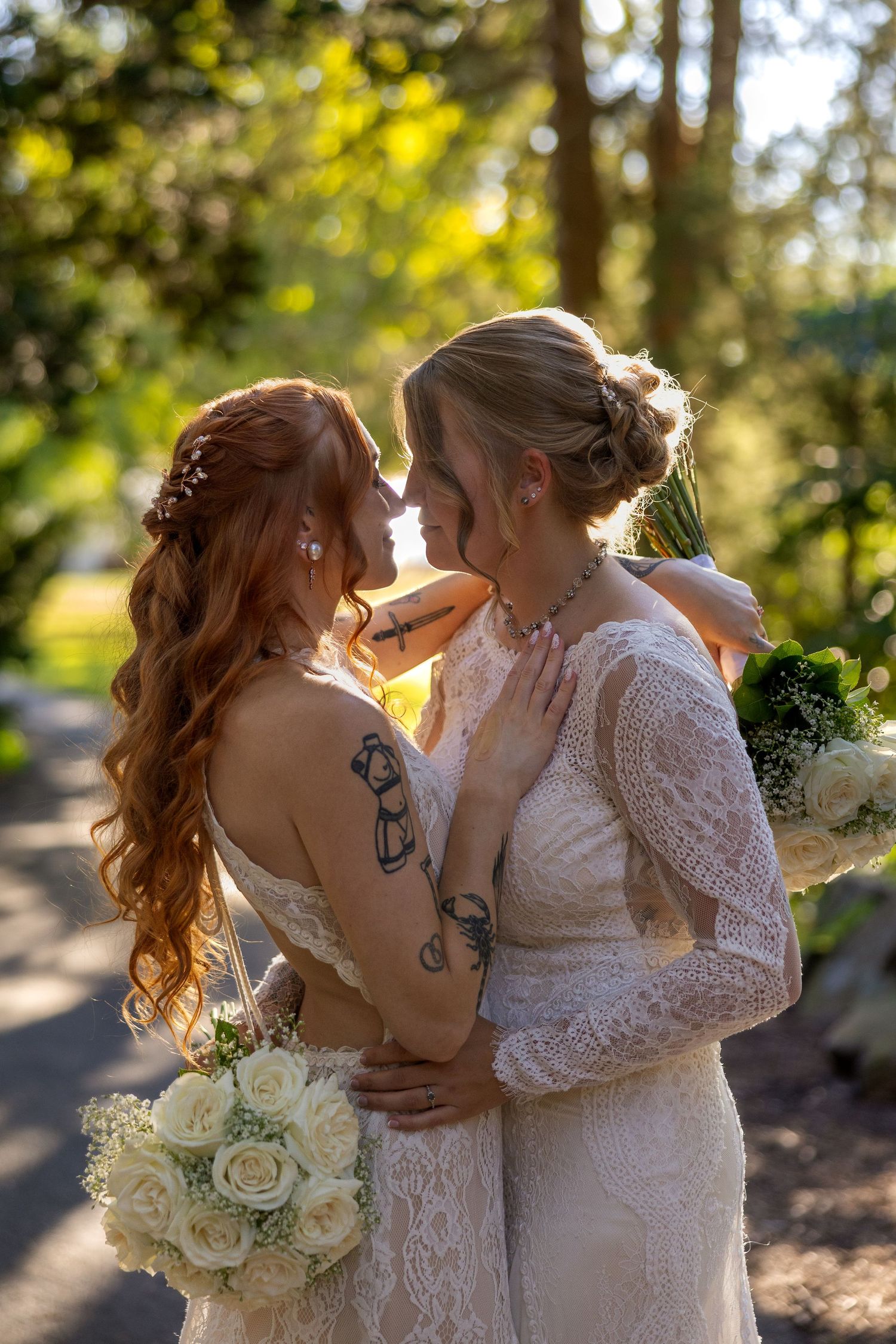 Two people in white lace wedding dresses share an intimate moment in a sunlit outdoor setting with white roses.