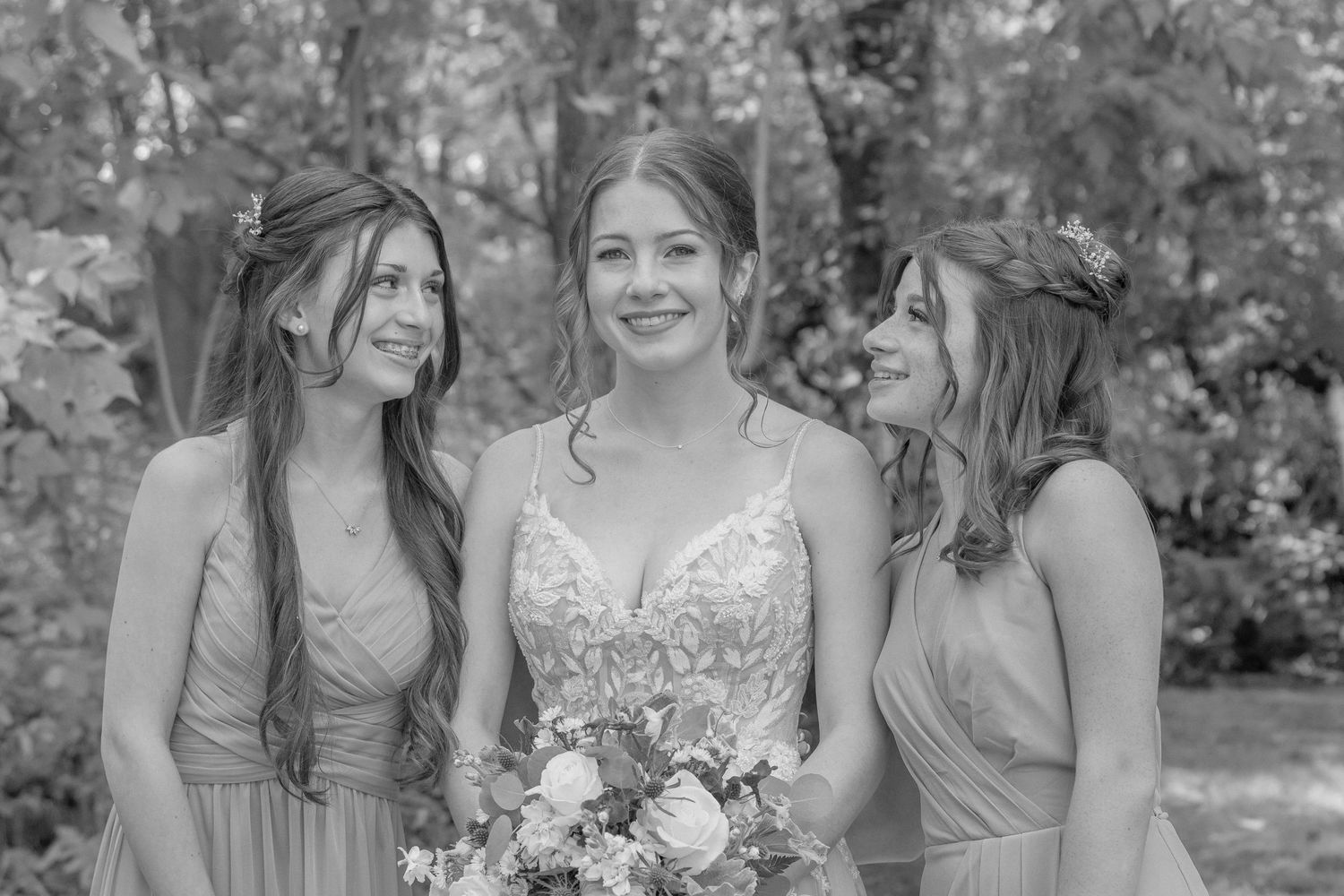 Bride in lace gown poses with bridesmaids holding floral bouquets in outdoor garden setting.