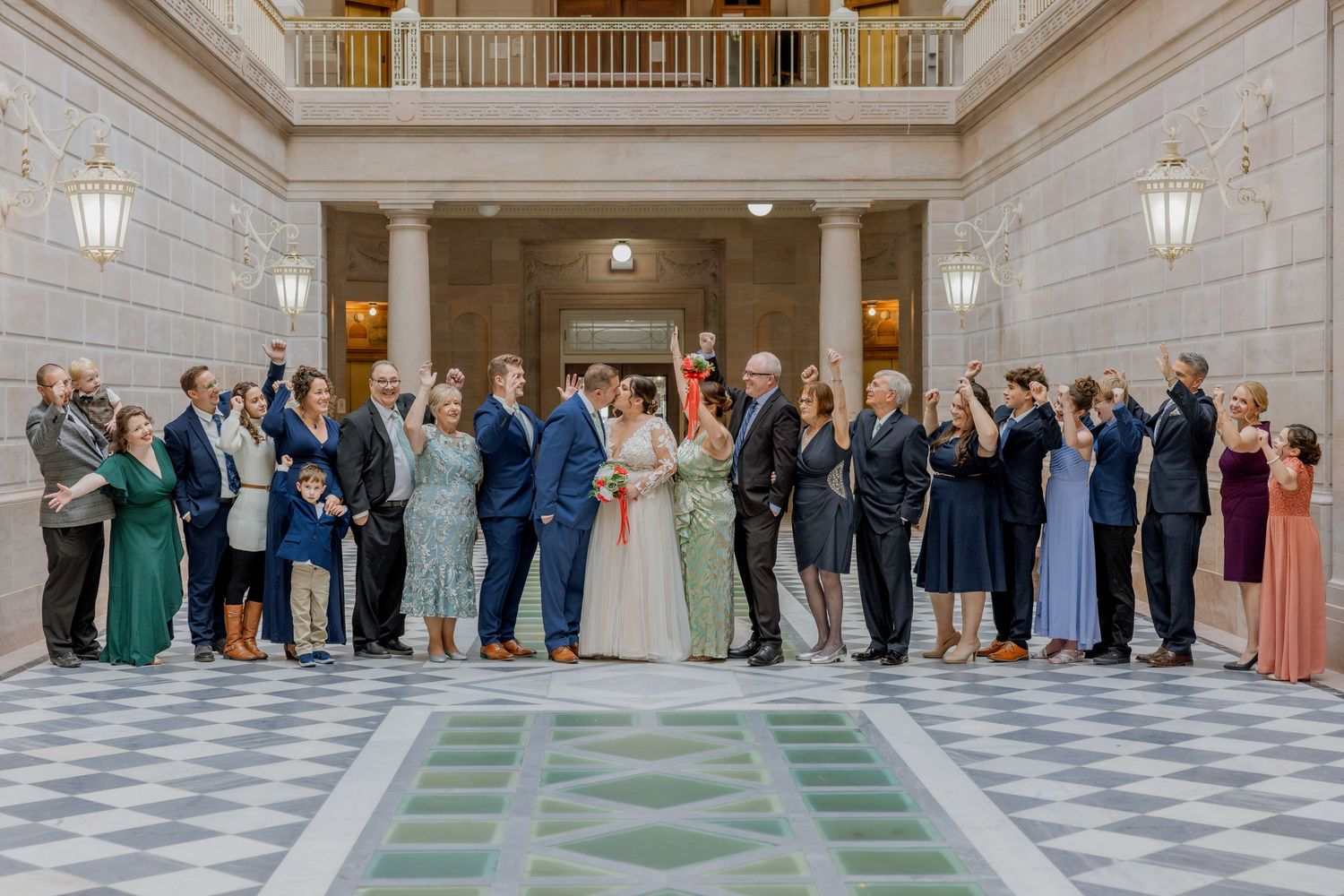 Wedding party lines up for group photo in ornate marble hall with checkered floor.
