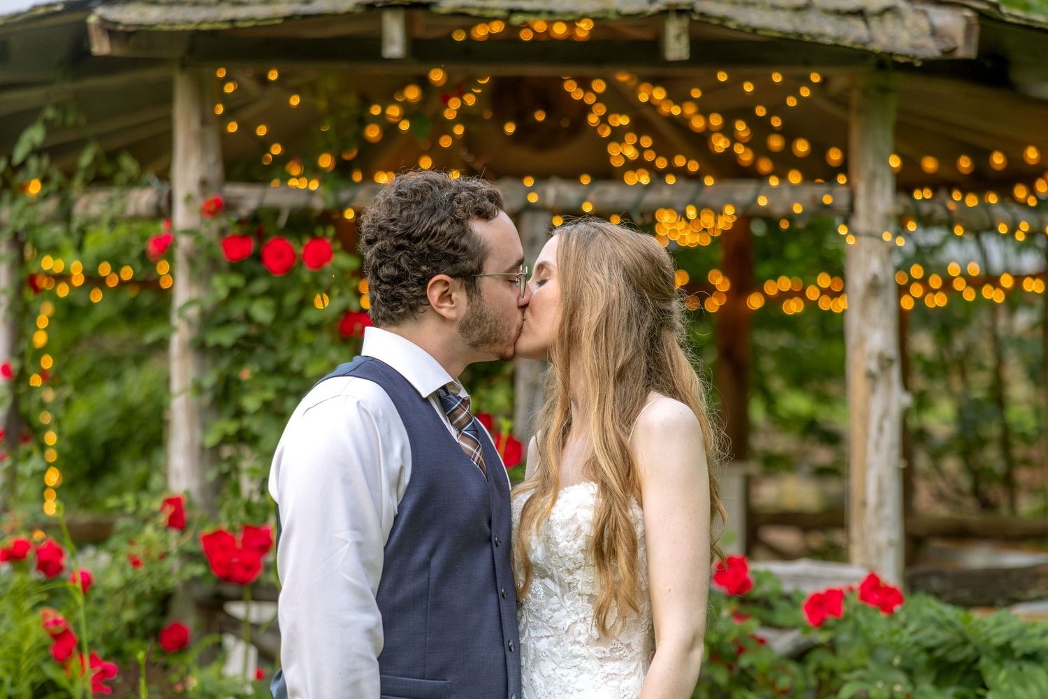 A couple shares a romantic kiss in a garden setting with twinkling lights and red roses in bloom.