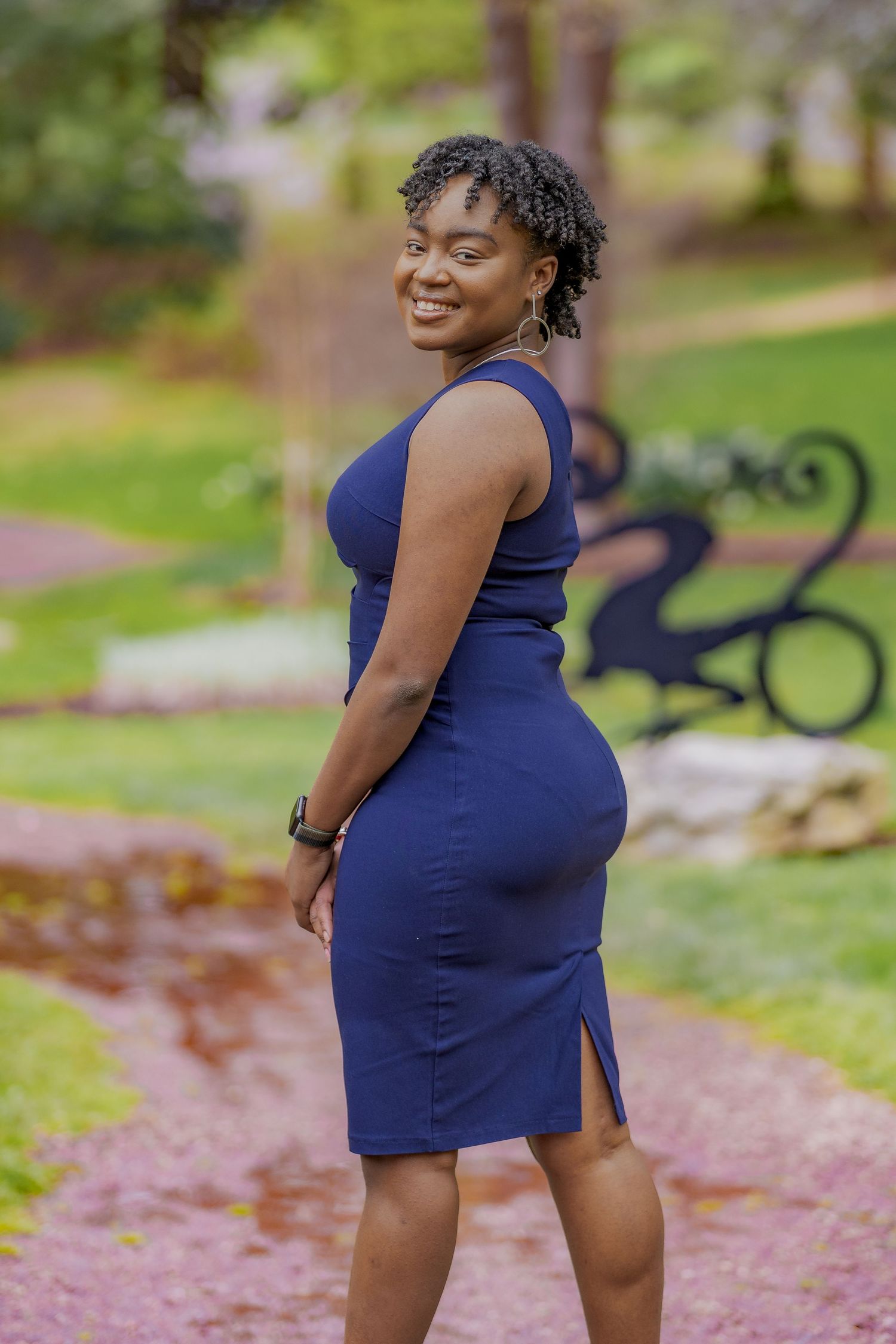 Person in fitted navy dress poses confidently on garden path with decorative bench in background.