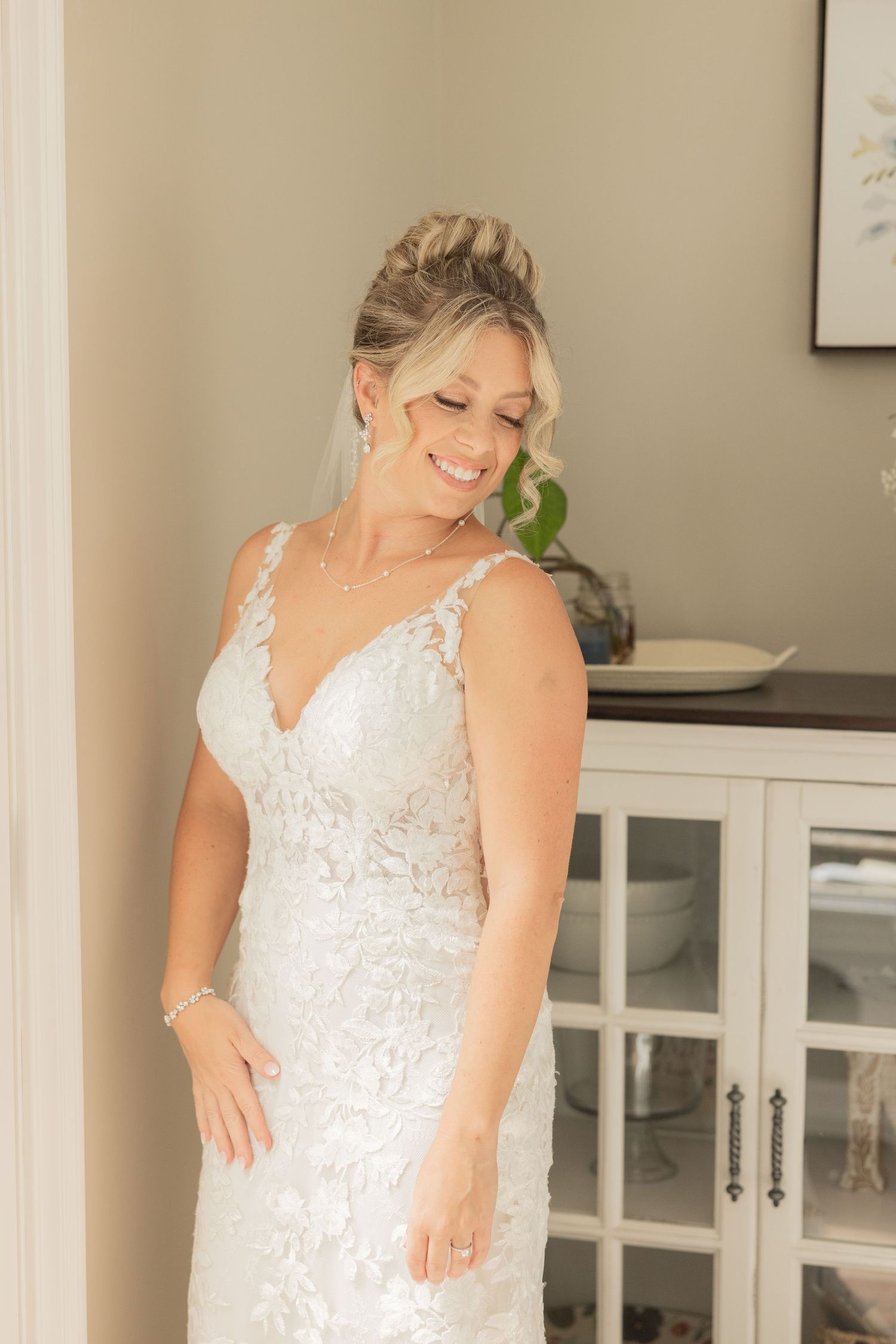Bride in lace wedding dress smiles warmly near white cabinet.