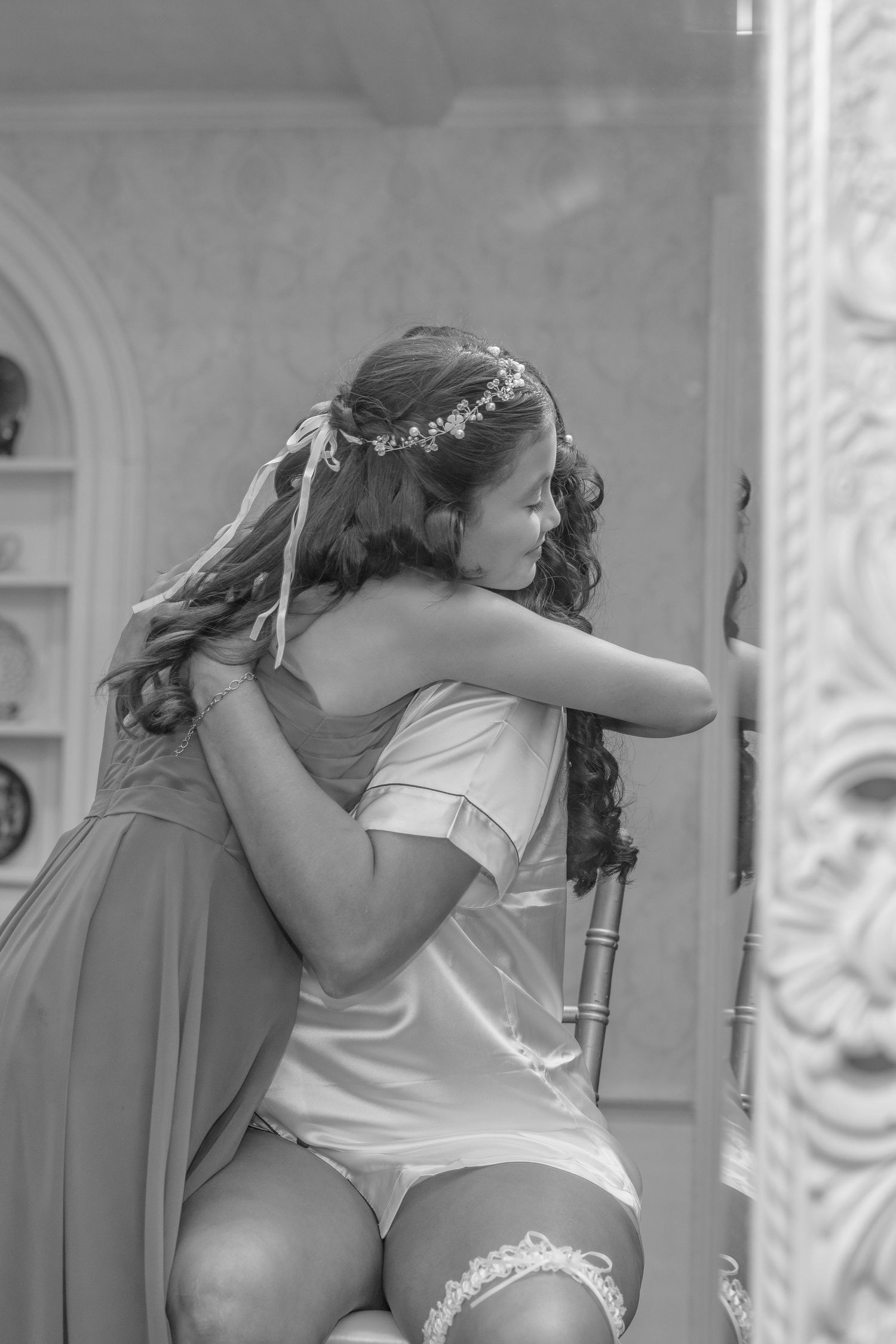Bridesmaid in flowing dress hugs bride during wedding preparation.