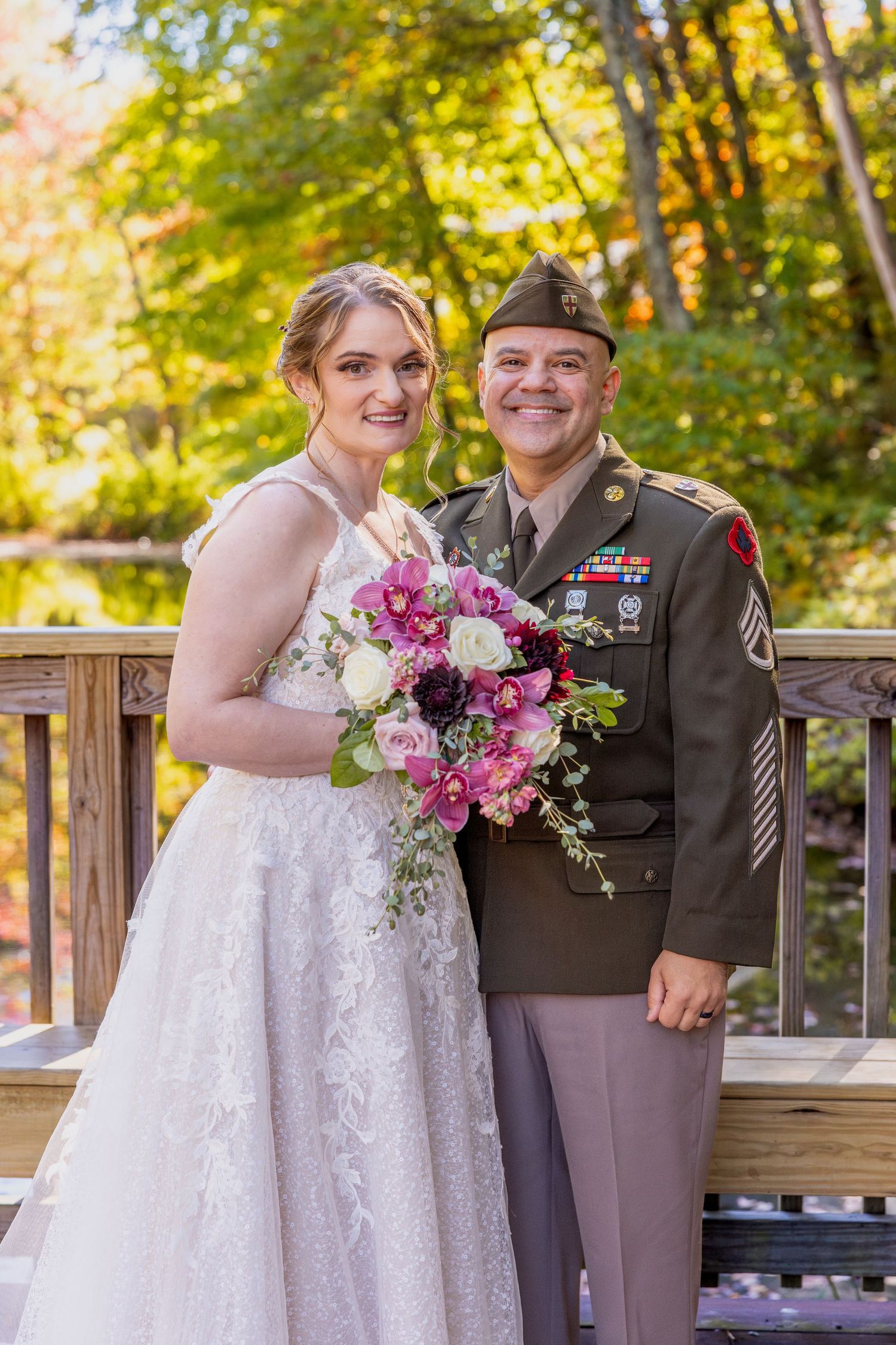 A bride in white lace gown poses with a servicemember in dress uniform holding pink flowers.