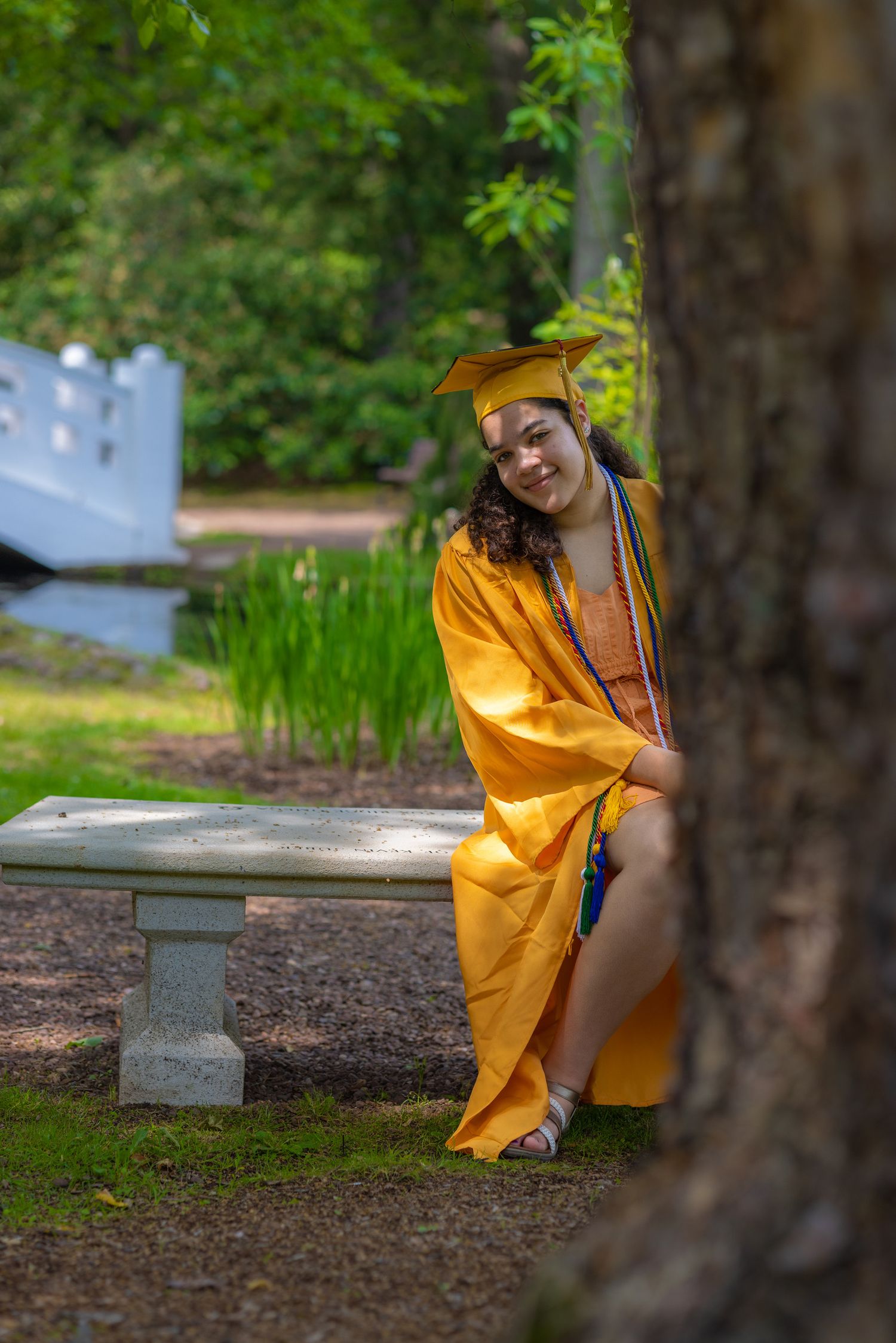Graduate in yellow academic regalia sits casually on stone bench in park setting.