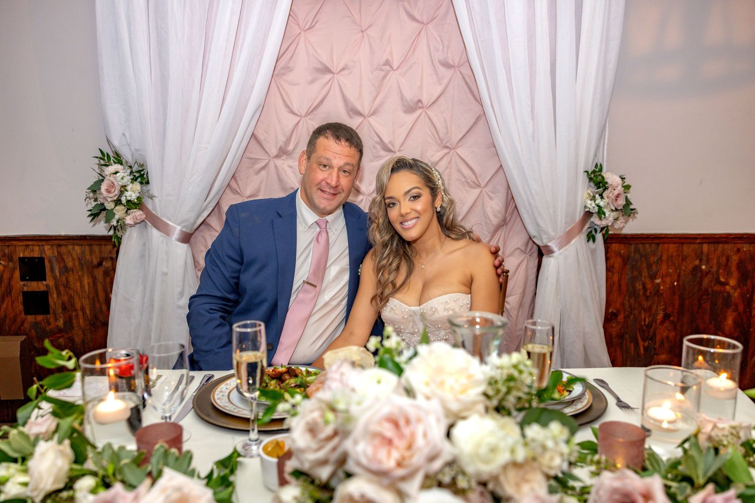 Elegant wedding reception table setting with pink roses and white floral centerpieces under draped fabric decor.