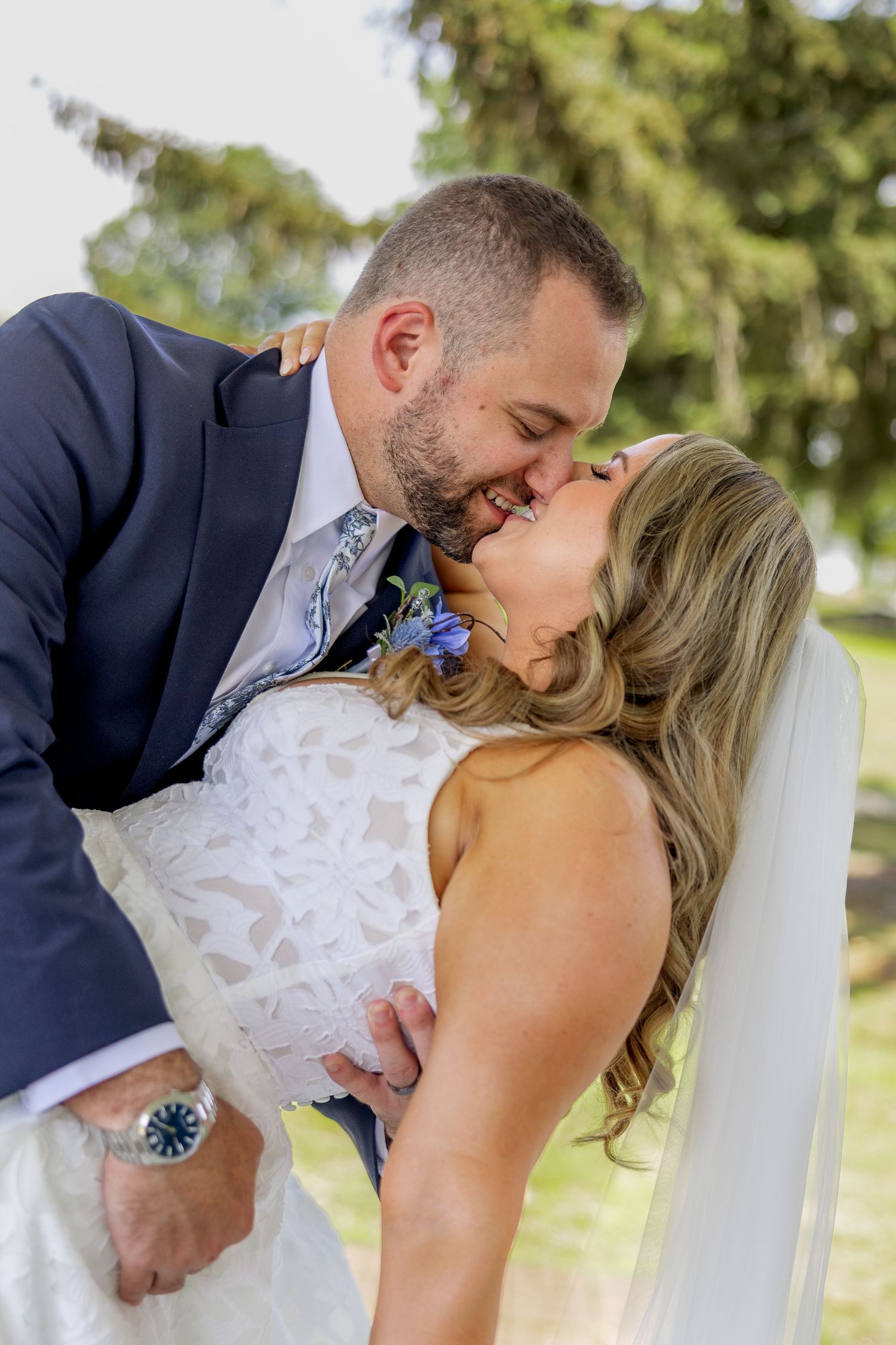 A couple in wedding attire shares a romantic kiss outdoors, with the bride in white lace and groom in navy suit.