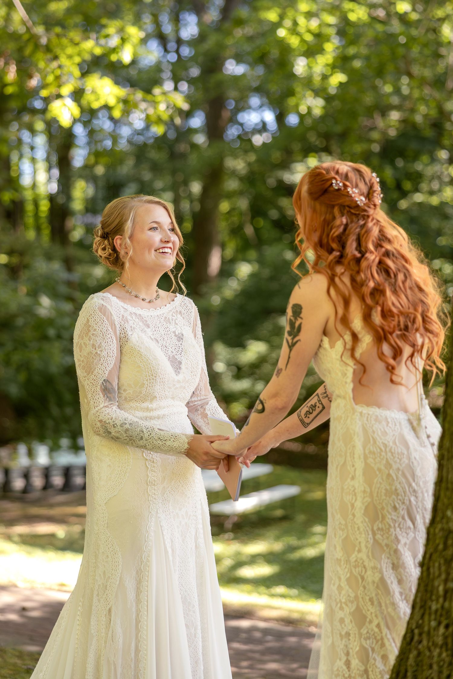 Two people in white lace wedding dresses share a joyful moment together in a sunlit forest setting.