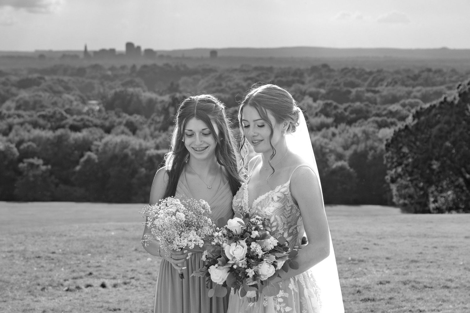Two people holding floral bouquets pose together on a hilltop with a scenic landscape view.