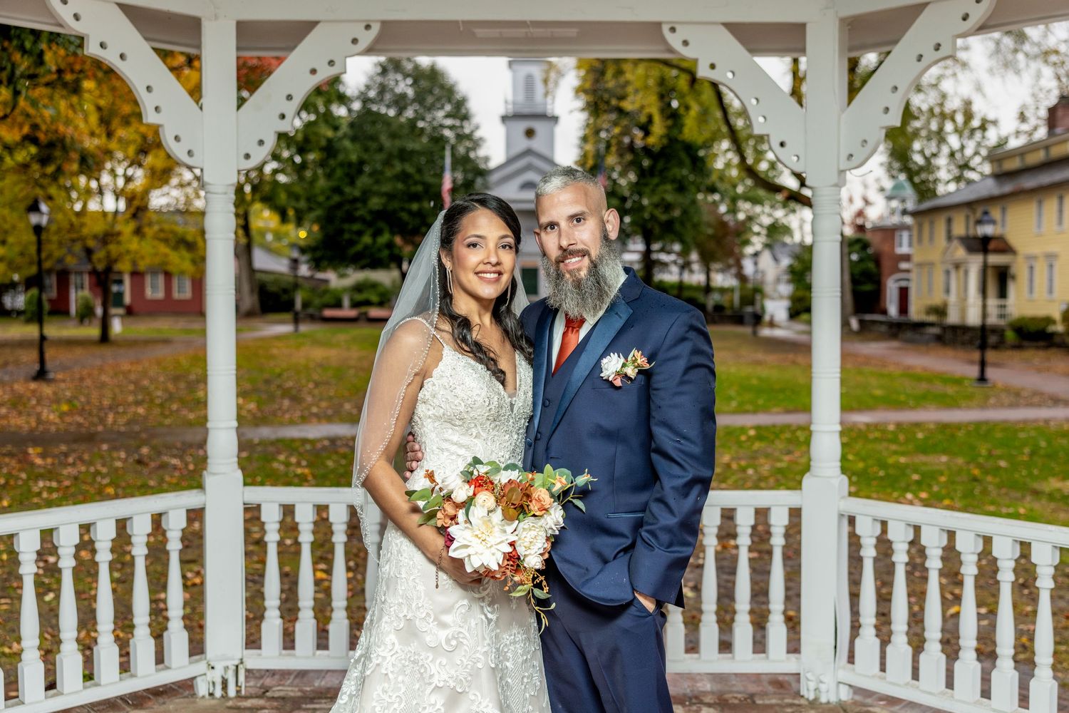 Wedding couple poses in white gazebo with church steeple and autumn foliage in background.