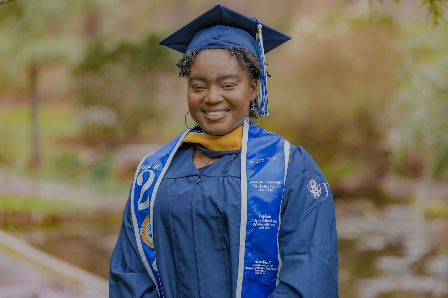 Graduate wearing blue academic regalia and honors cords smiles during outdoor ceremony.
