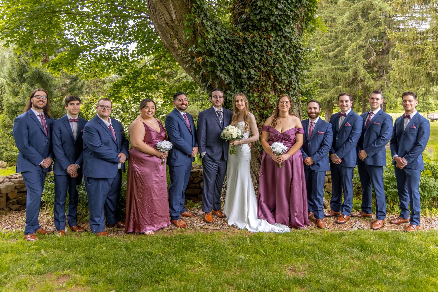 Wedding party in mauve dresses and navy suits poses outdoors in a garden setting.