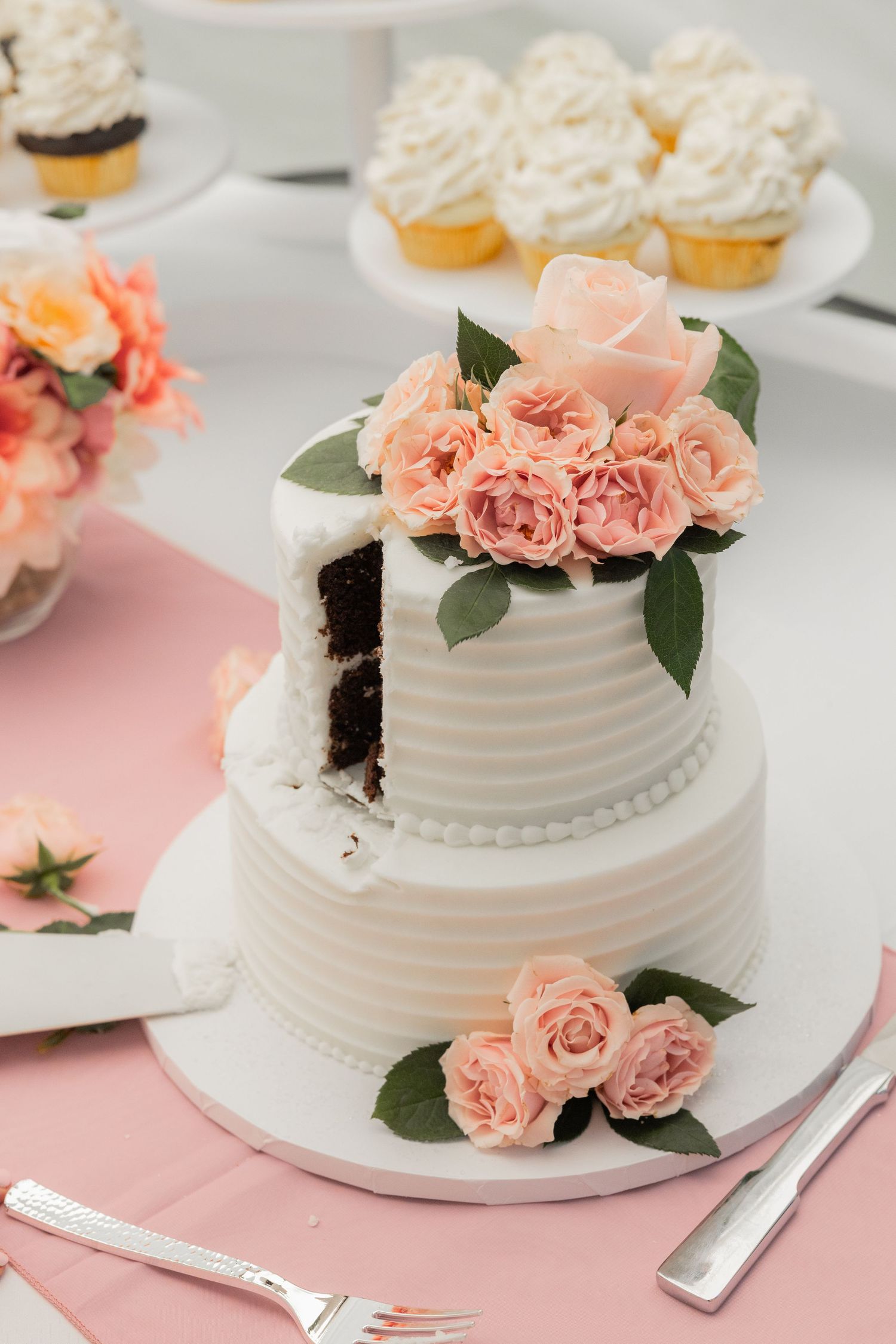 A two-tiered white wedding cake decorated with peach roses and greenery sits on a pink tablecloth.