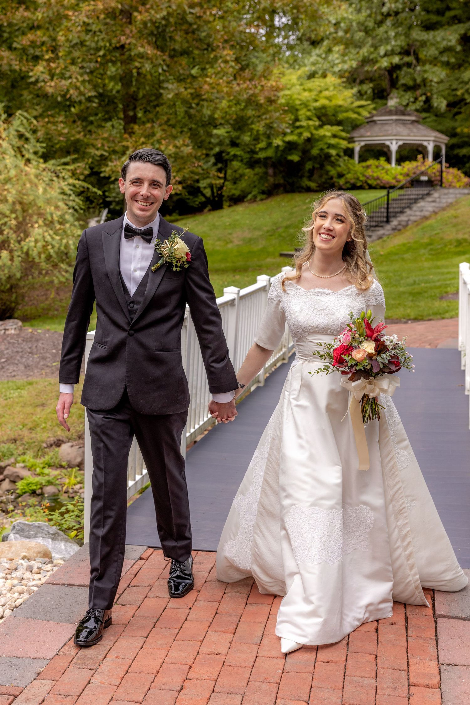 Joyful wedding couple walks hand in hand on brick pathway with gazebo in background.