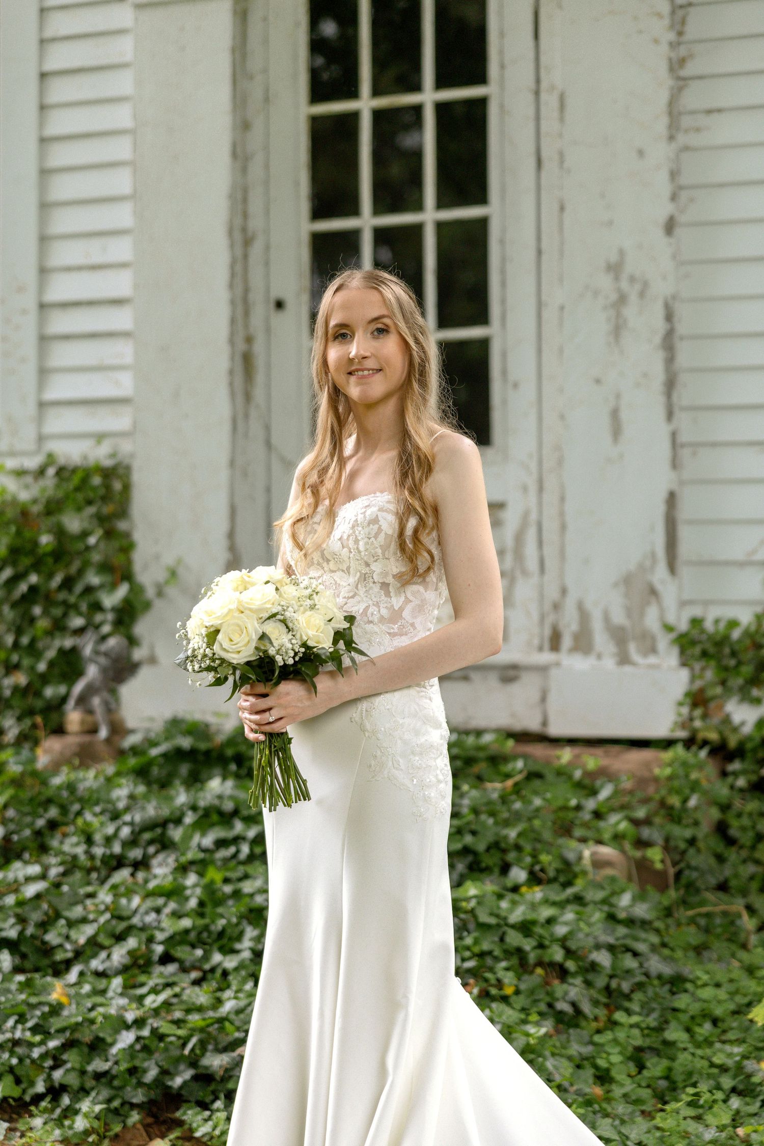 Bride in white strapless gown poses with bouquet of white roses against rustic white siding and greenery backdrop.
