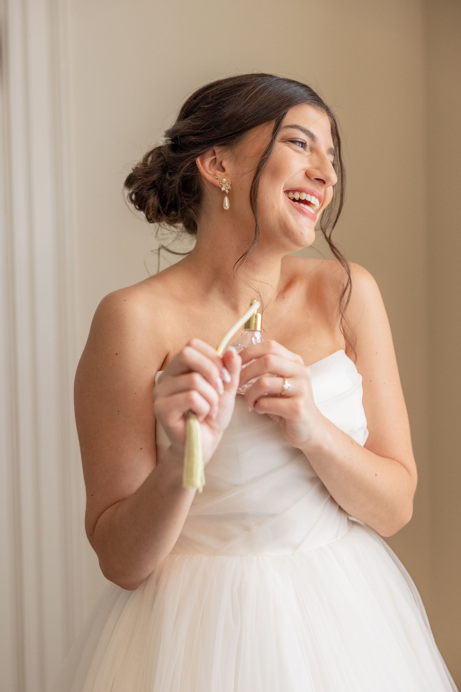 Joyful bride in white dress laughs while holding something near window.