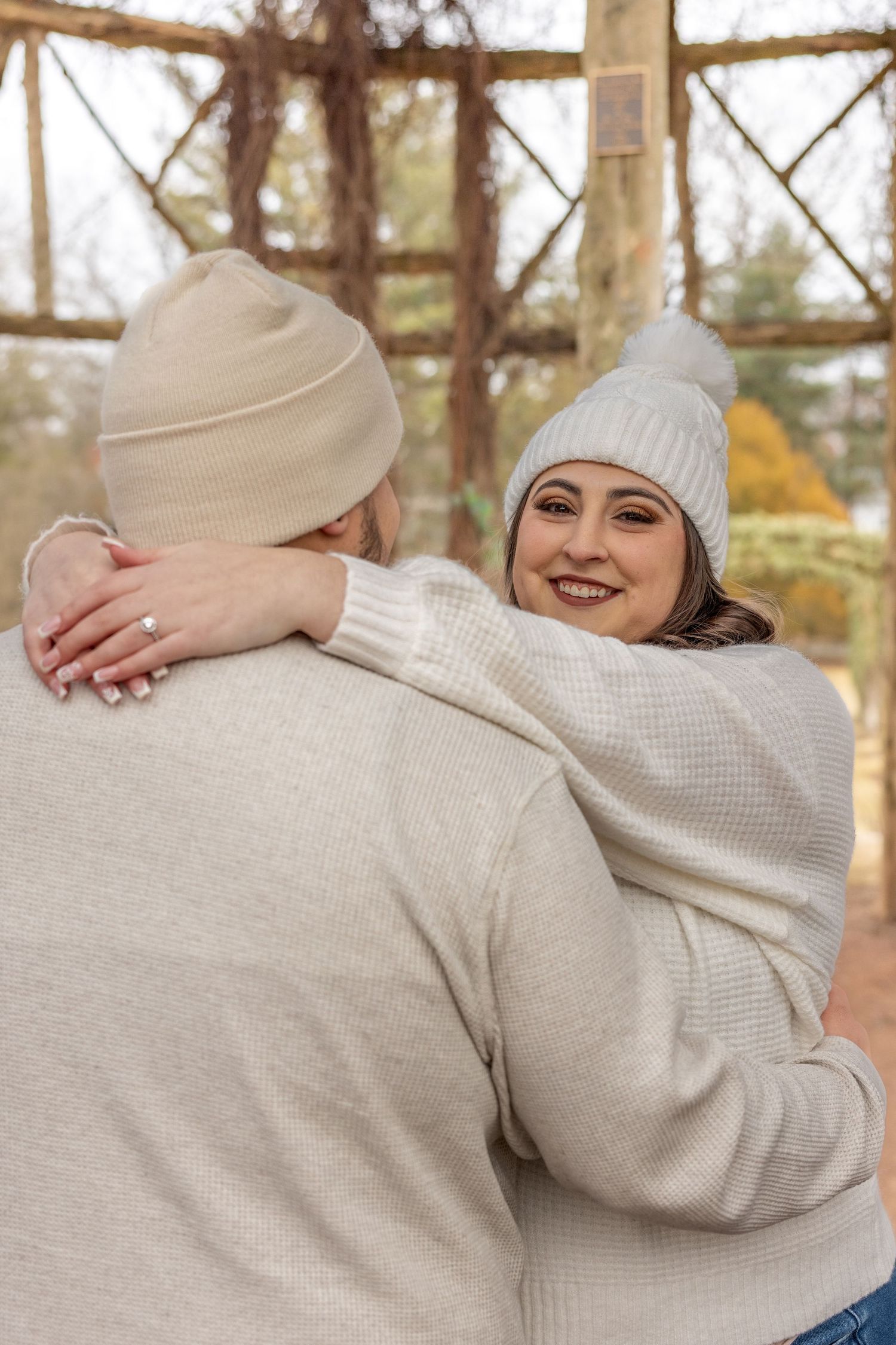 A couple in matching white sweaters and winter hats embraces on a rustic wooden porch.