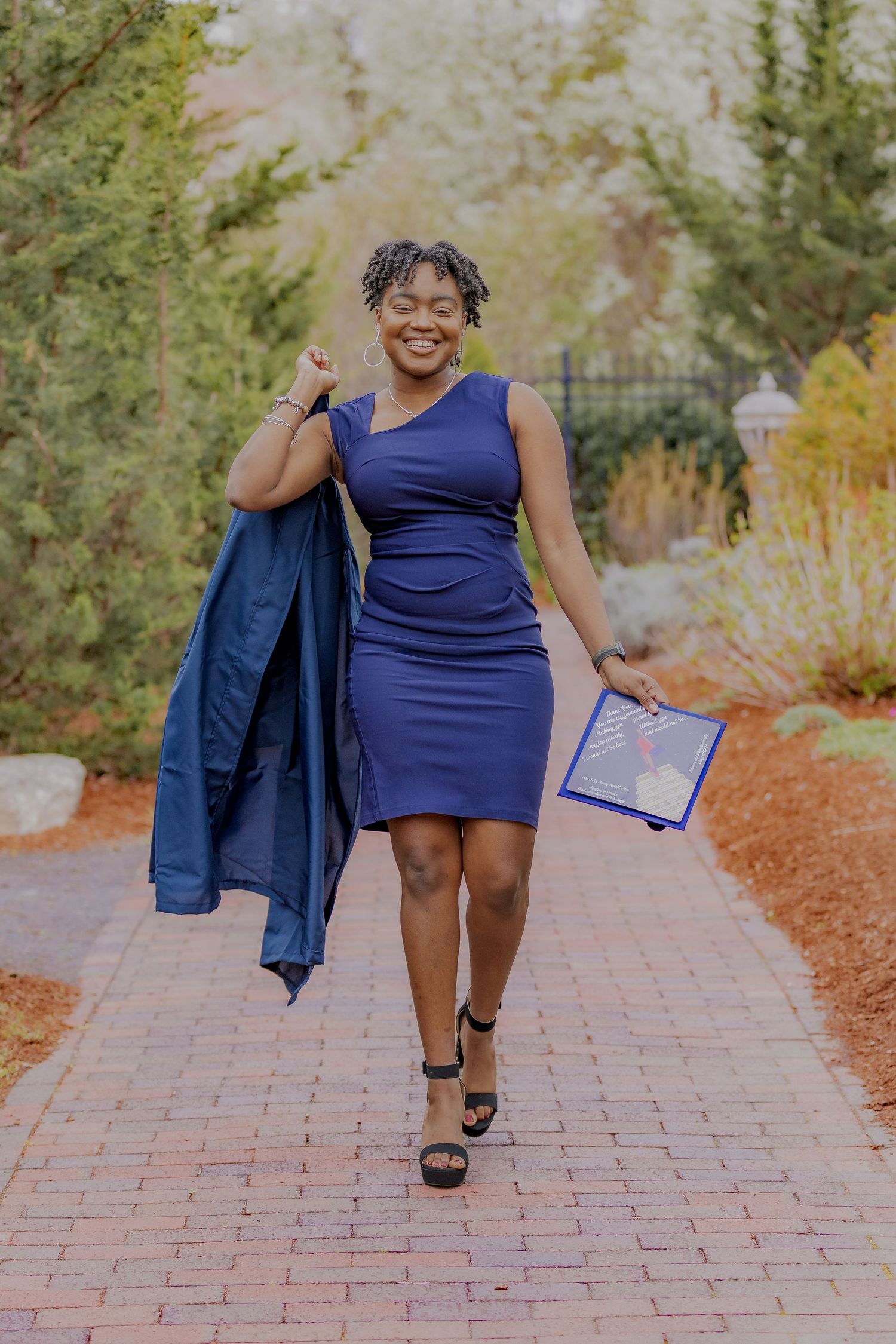 A person in a navy blue dress walks along a brick path carrying a jacket and folder on an autumn day.