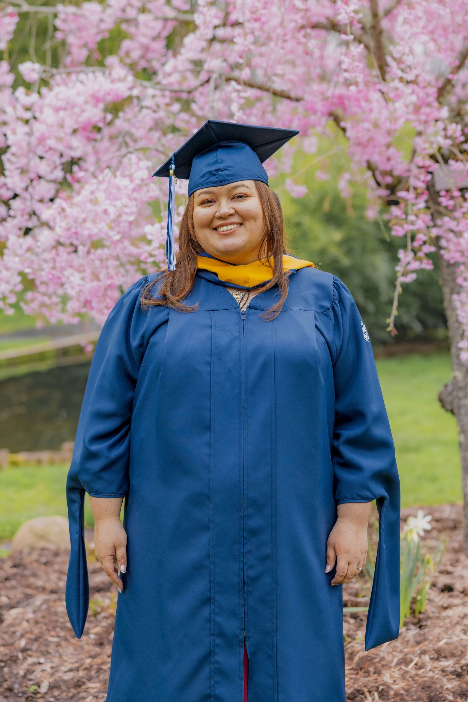 Graduate posing in blue cap and gown with cherry blossoms blooming in background.