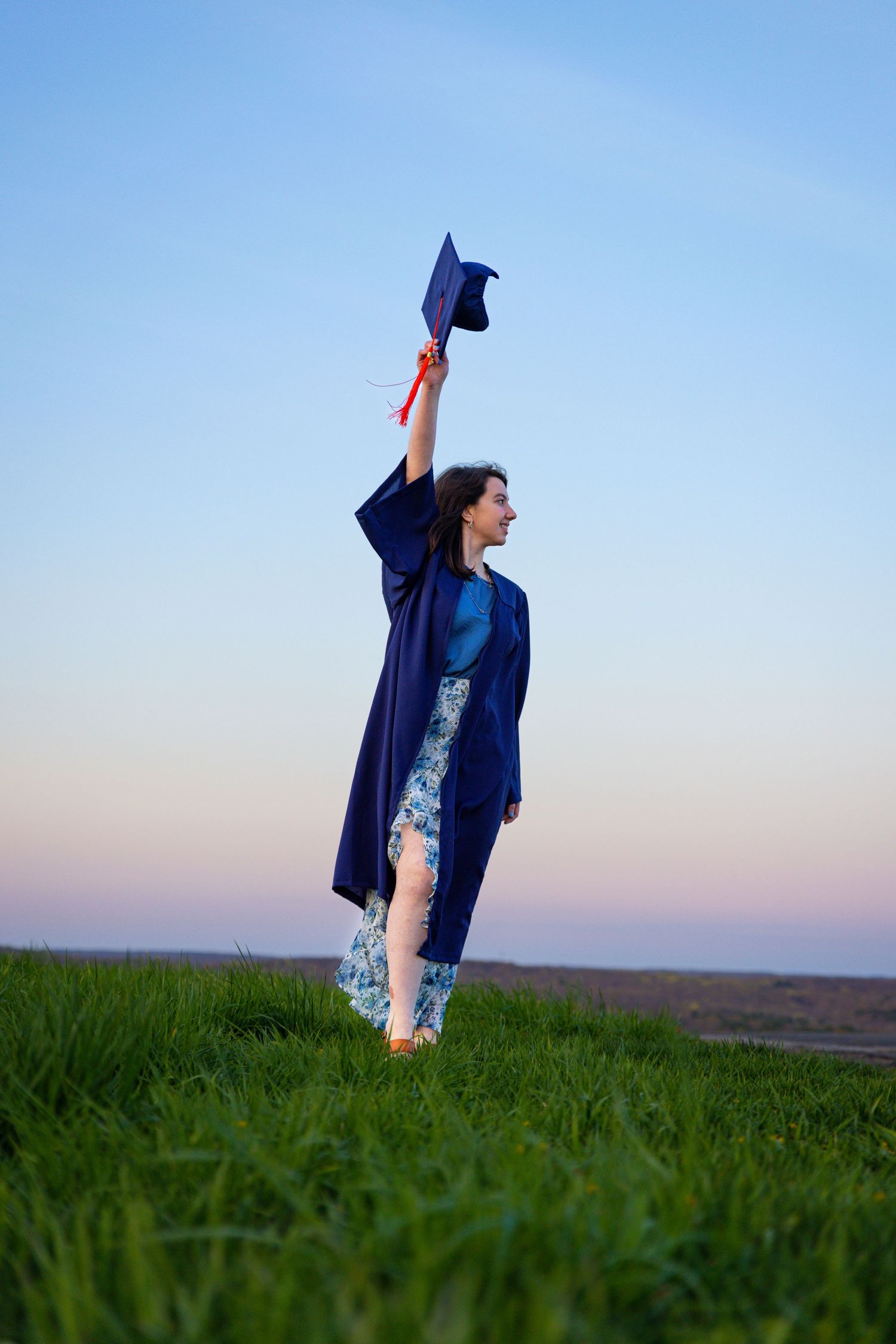 Graduate in blue cap and gown throws mortarboard in air while standing in grassy field at sunset.