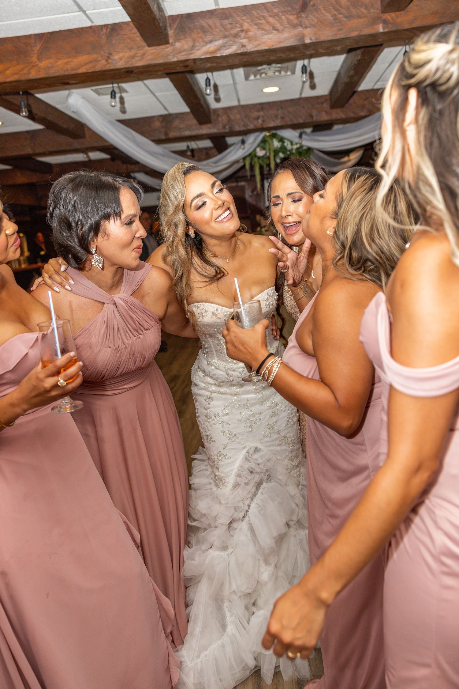 Bridesmaids in dusty rose dresses celebrate joyfully with the bride at a rustic indoor wedding venue.