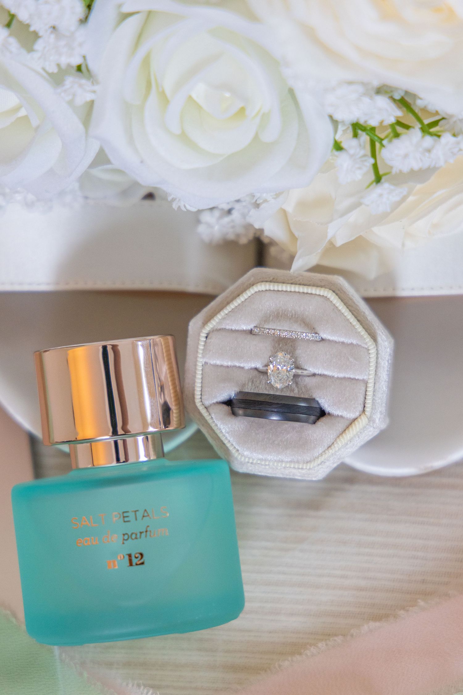Wedding rings and engagement ring displayed with white flowers and teal jewelry boxes against a light wood surface.
