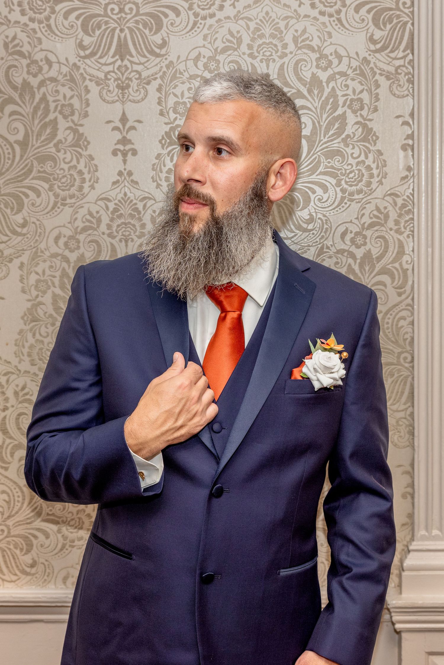 Groom in navy suit with orange tie poses against ornate wallpaper background.