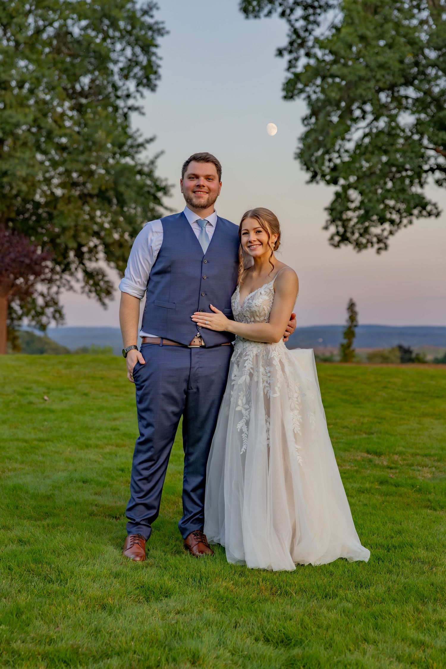 Sunset wedding portrait with couple standing in meadow with moon visible in sky.