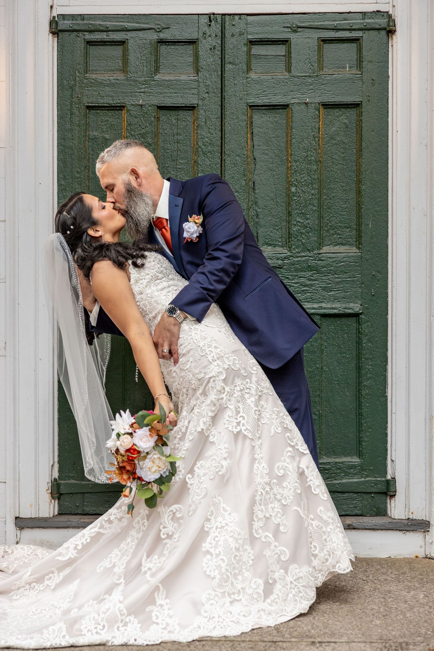 Couple shares romantic kiss against vintage green door in formal wedding attire.