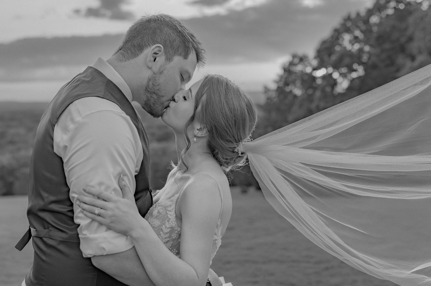 A black and white wedding portrait with a flowing veil caught in the wind at dusk.